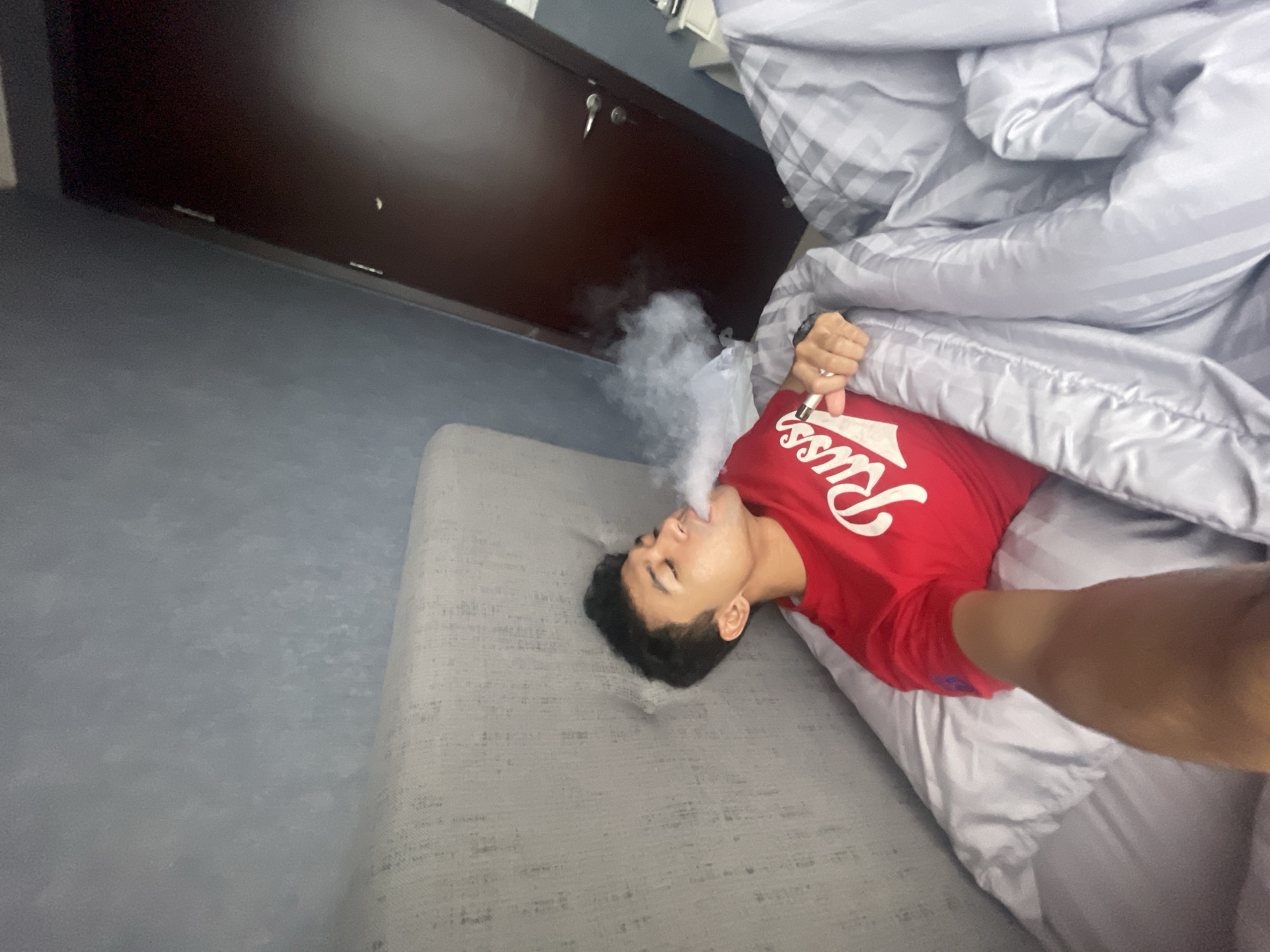 A man is lying on a gray bed with a gray and white striped comforter in a room with gray walls and a dark brown cabinet. He is wearing a red t-shirt and is exhaling smoke from a device in his hand. The location is Central Jakarta, Indonesia.