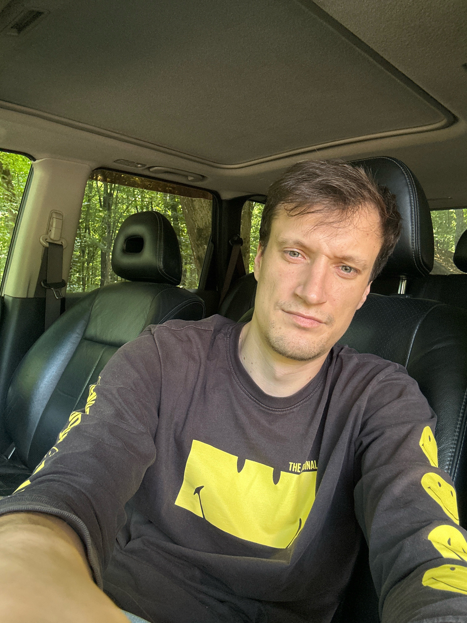 A man with short, brown hair and a slight stubble is seated in the driver's seat of a car, looking directly at the camera. He is wearing a dark, long-sleeved shirt with a large yellow smiley face graphic on the front and smaller smiley faces on the sleeves. The car's interior is visible, including black leather seats and a sunroof, which is partially open. Through the windows, a lush, green forest can be seen, suggesting the car is parked in a wooded area. The overall lighting indicates it is daytime, with natural light filtering into the vehicle. The specific location within Shatava, Ukraine cannot be determined from the visual information present.