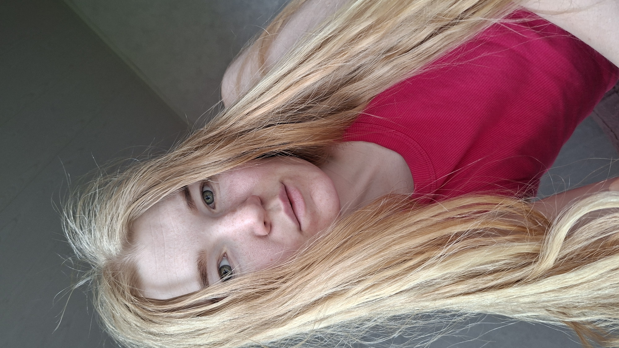 A headshot of a fair-skinned person, likely female, is shown lying down and looking directly at the camera. The image is rotated, presenting the person's face on the left side of the frame, with their body extending to the right. The subject has long, straight, blonde hair that spreads around their head and shoulders, displaying varying shades of blonde. Their eyes are light-colored, appearing greenish-grey, and they have a neutral, soft expression. The skin shows slight redness on the cheeks and nose, and a few freckles are visible. The person is wearing a bright red top with a ribbed texture, partially visible underneath the hair. The background is a plain, light grey surface, suggesting an indoor setting. No specific activity beyond posing for the photograph is evident. There is no visible text in the image. The location context for this image is Большой Букор, Russia.