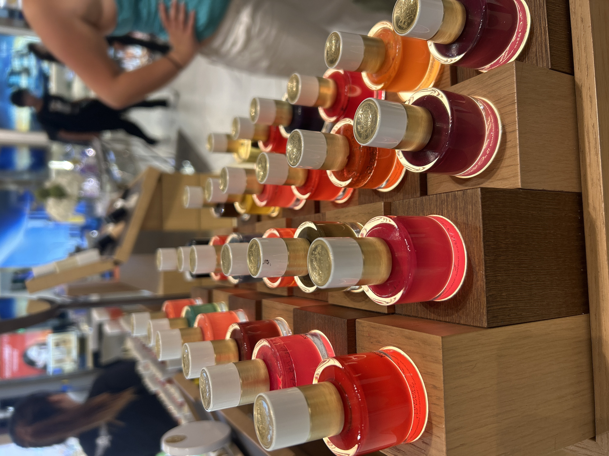 In Tambon Mai Khao, Thailand, a brightly lit retail store displays an array of perfume bottles. The perfumes are arranged in tiered wooden stands, with each bottle featuring a cylindrical glass base filled with colored liquid and topped with a white and gold cap. The colors of the perfumes range from vibrant reds and oranges to deep purples and greens. In the blurred background, several individuals are visible. To the left, a person with dark hair pulled back in a ponytail is wearing a black shirt. In the center, another individual in a black t-shirt and pants is walking away from the camera. On the right, a person in a turquoise top and white skirt is facing away from the viewer, with their right hand on their hip. The store environment appears to be a beauty or fragrance shop, with shelves and displays visible in the background.