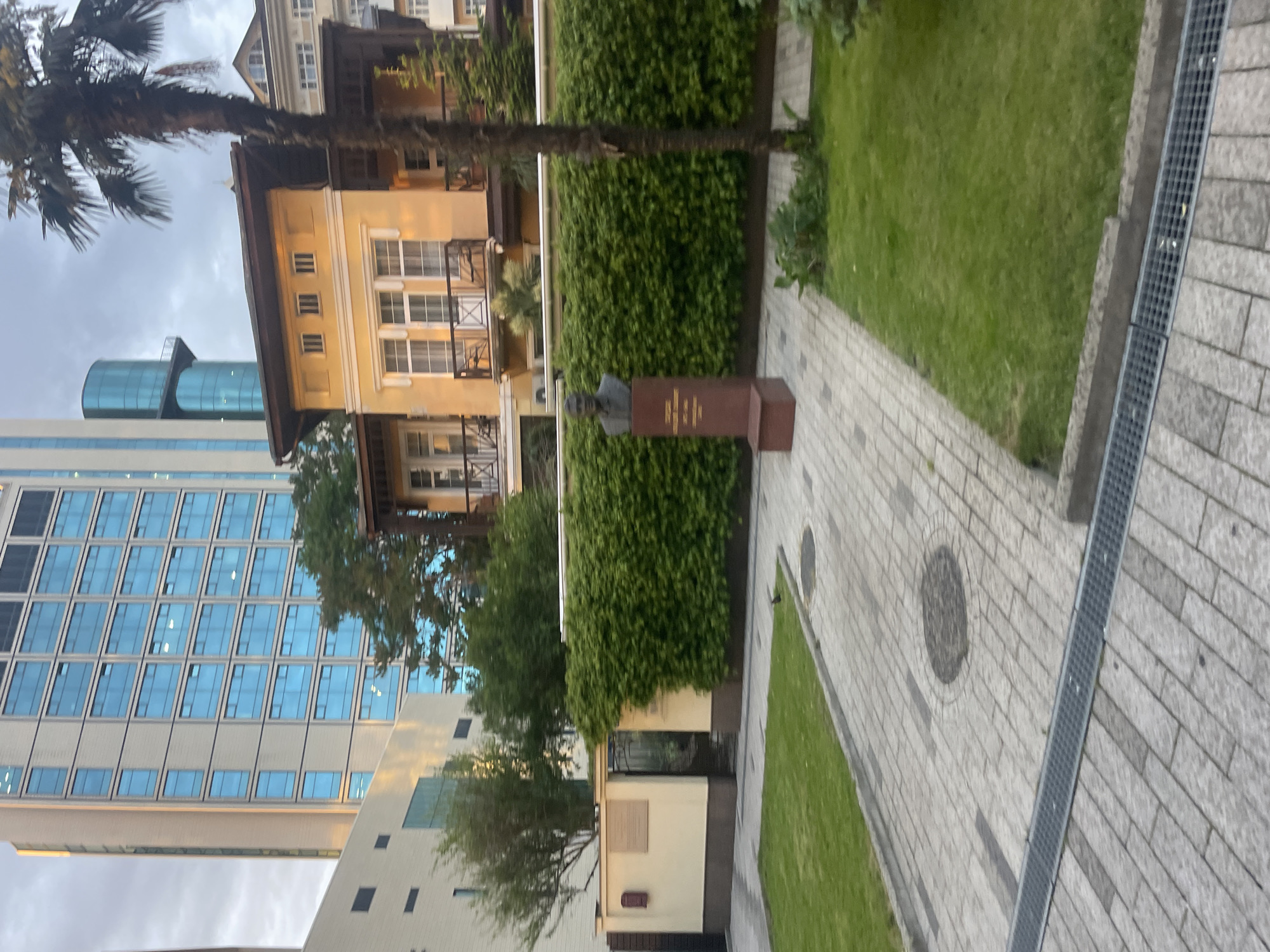 The image shows a street scene in Sochi, Russia, with a view of modern buildings and older architectural styles. A statue rests on a pedestal behind a green hedge on a paved walkway alongside a strip of grass. The building to the left is a modern high-rise with glass windows. To the right and partially obscured, there's a yellow multi-story building with balconies, possibly an older residential building. A palm tree is visible in the background. The sky appears overcast.