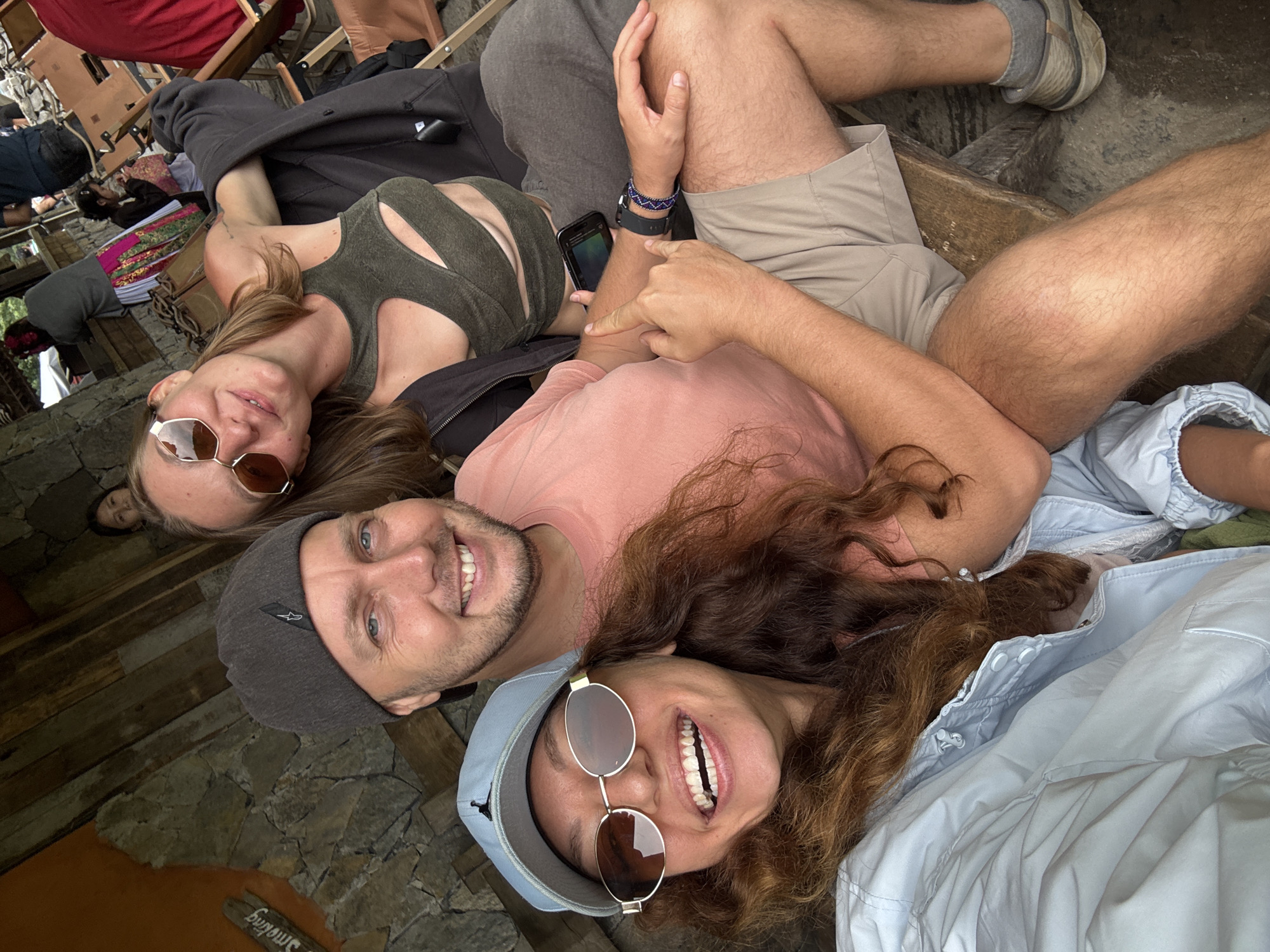 Three people are lying on a stone and wooden structure in Phố Bảng Commune, Vietnam, based on the sign that says "Bảng" visible in the image. The person on the left has on sunglasses and a green top. The person in the middle has on a pink shirt and a grey beanie. The person on the right has on sunglasses and a blue baseball cap and is wearing a blue jacket. Other people are in the background.