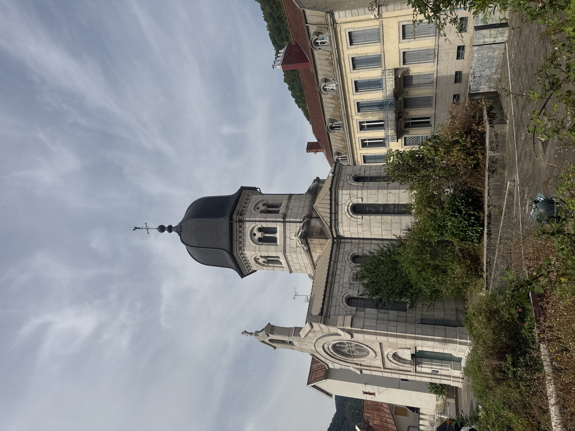 The image shows a view of the Saint-Pierre Cathedral in Saint-Claude, France. The cathedral is a stone building with a dark dome and a cross on top. To the right of the cathedral is a yellow building with a balcony and several windows. The sky is cloudy with some patches of blue. The foreground of the image shows some overgrown vegetation and a concrete pathway.