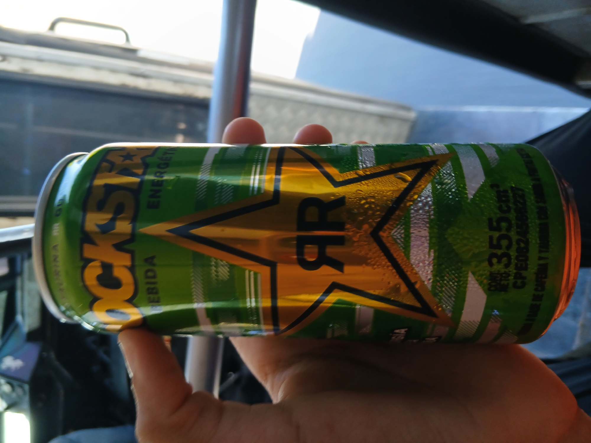 A hand holds a green and gold Rockstar energy drink can. The can is cylindrical and features the Rockstar logo prominently. The can has condensation on the surface. The background shows a vehicle interior with parts of the window, door, and metal bar visible. Text on the can indicates it contains 355cm³ and is based on caffeine and taurine. The location in Altagracia, Venezuela is not deducible from the image.