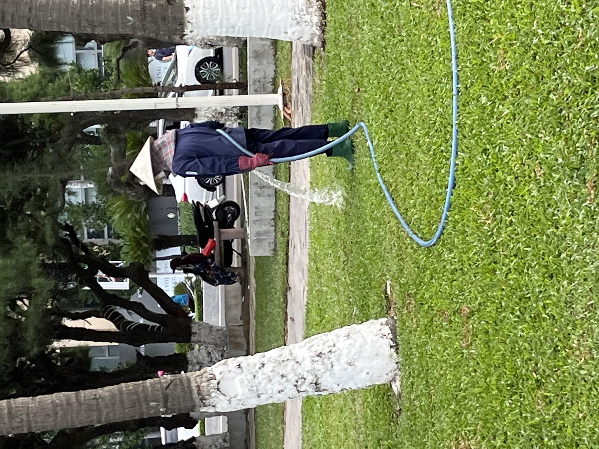 A person wearing a conical hat, a blue uniform, pink gloves, and green boots is watering grass with a blue hose. The person is standing in a park-like setting with trees and a white-painted tree trunk in the foreground. In the background, there are buildings, a street with cars and a motorcycle, and two people sitting on a bench. The scene appears to be in Nha Trang, Vietnam, a coastal city known for its beaches and parks.