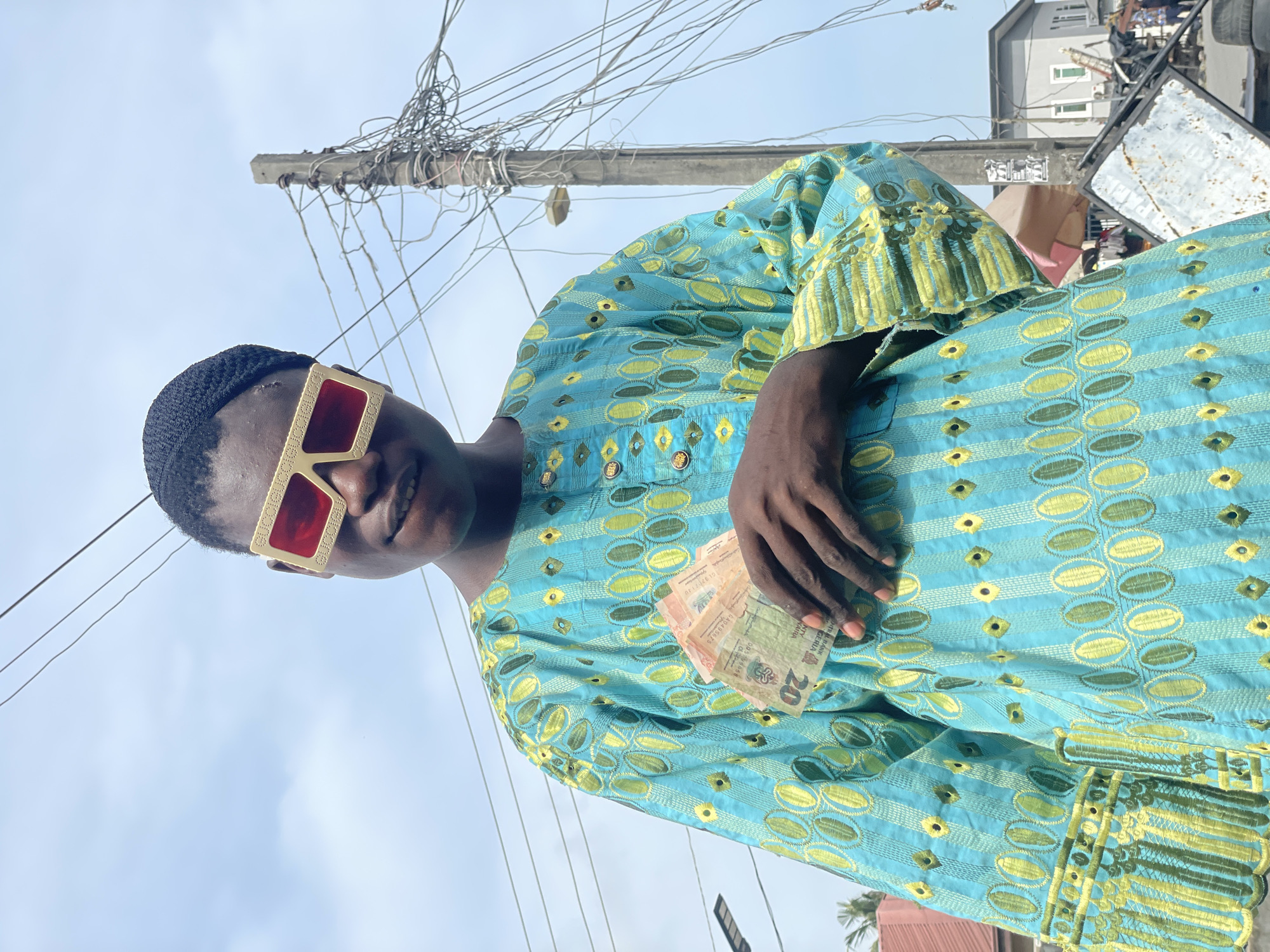 The image shows a person wearing a blue and yellow patterned garment, a black cap, and red-tinted sunglasses. The person is holding a stack of Nigerian Naira notes in their left hand. Behind the person is a utility pole with many wires, and a building with windows can be seen in the background. The scene appears to be outdoors under a bright sky. The person is smiling at the camera. The Nigerian Naira notes have the number 20 printed on them.