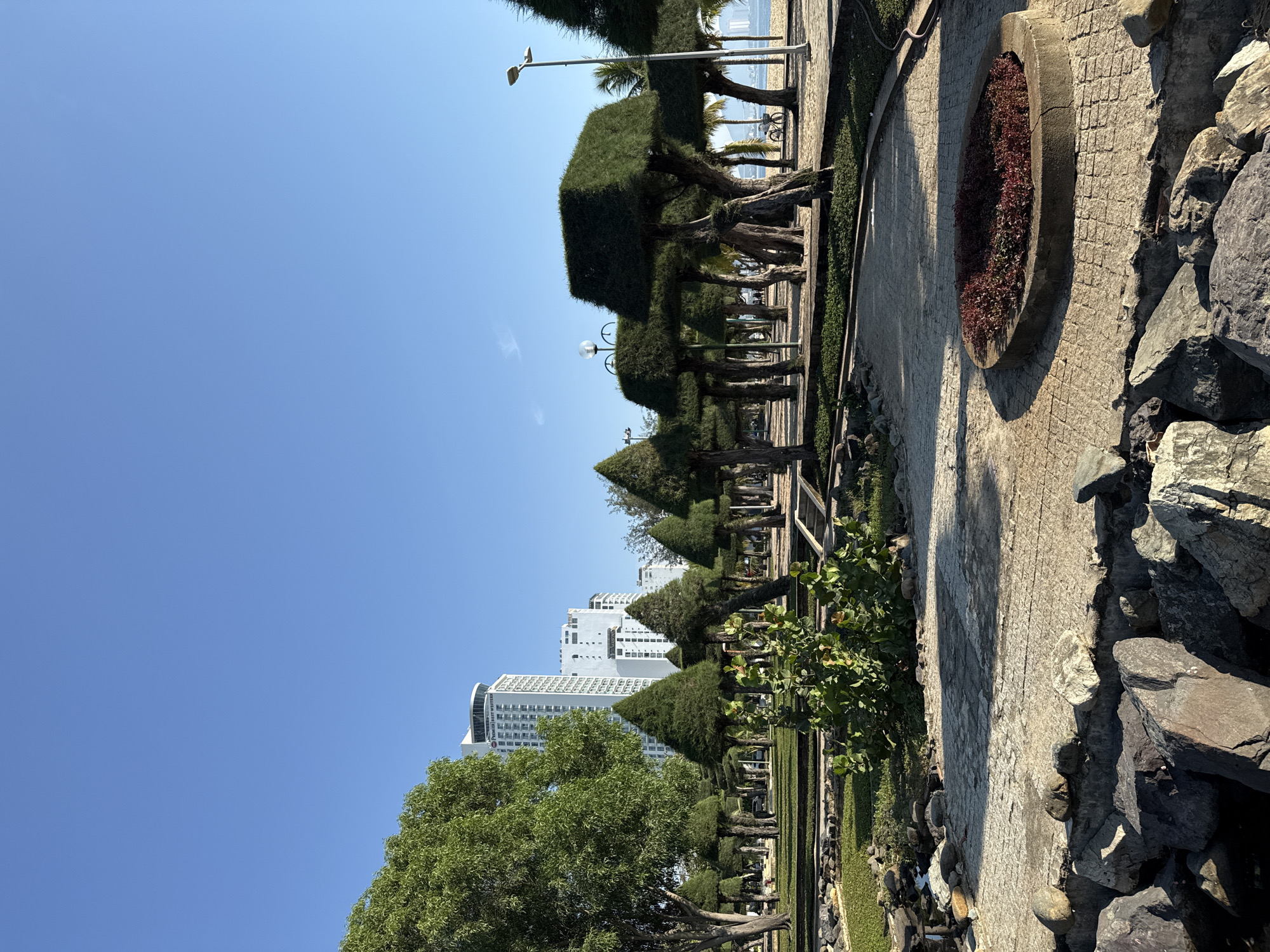 The image shows a park in Nha Trang, Vietnam, with the ocean and high-rise buildings in the background. The park features manicured trees, a paved walkway, and a circular flower bed. Several large rocks line the edge of the park, and a body of water is visible in the lower left corner. The sky is clear and blue, indicating a sunny day. There are no people visible in the image.