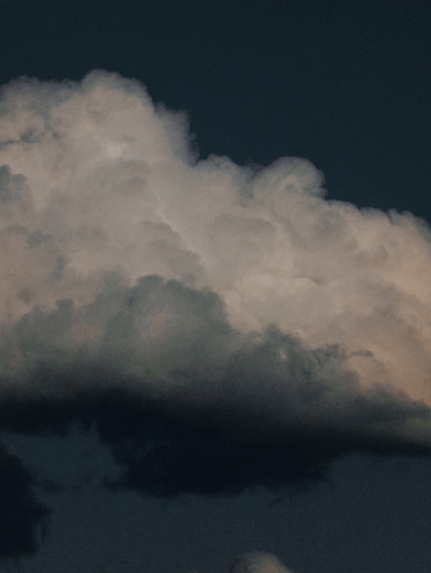 A close-up, low-angle shot captures a dramatic, dark sky filled with voluminous clouds. The clouds dominate the frame, exhibiting a gradient of tones from bright white and light pink near the top to deep grey and black at the bottom. The texture of the clouds is soft and wispy, with subtle undulations and shadows that suggest depth and movement. The overall lighting is dim, giving the scene a moody and somewhat ominous atmosphere. The image is devoid of any human presence or man-made structures.
