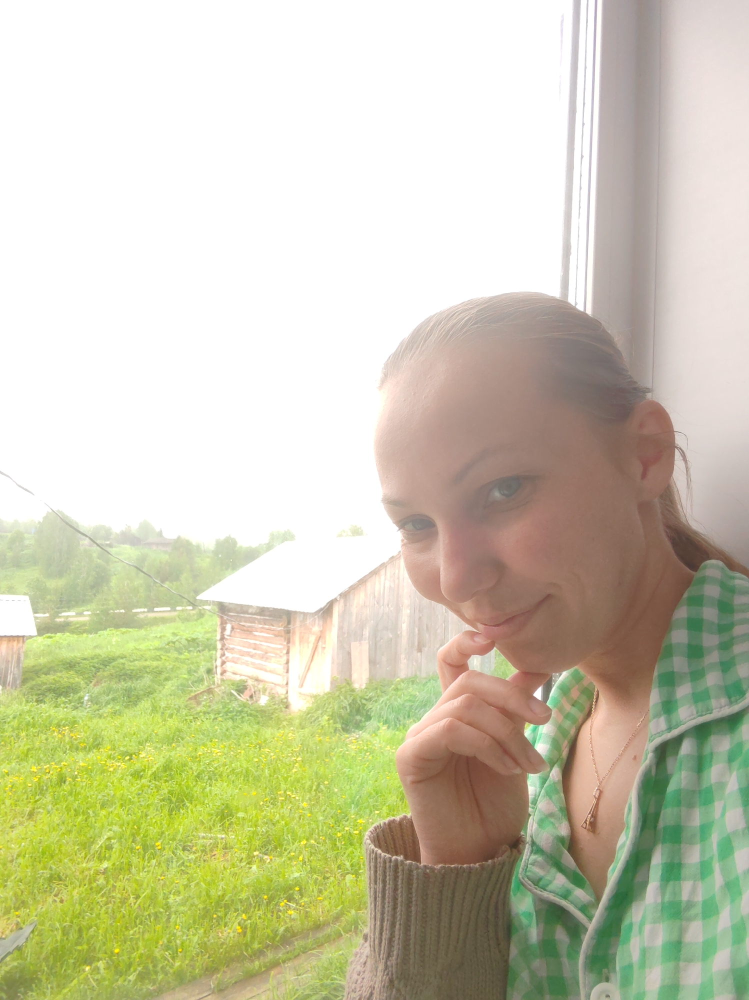 A woman is positioned in front of a window, with a rural scene visible outside. The woman has fair skin, light eyes, and her hair is pulled back. She is wearing a green and white checkered shirt over a sweater. She has a necklace with a charm. Her left hand is raised to her chin.
Outside the window, the scene features a field of green grass interspersed with small yellow flowers. There is a wooden structure with a slanted roof. There are trees and vegetation in the background. It appears to be a cloudy day, with soft lighting.
The location is Чухлэм, Russia.