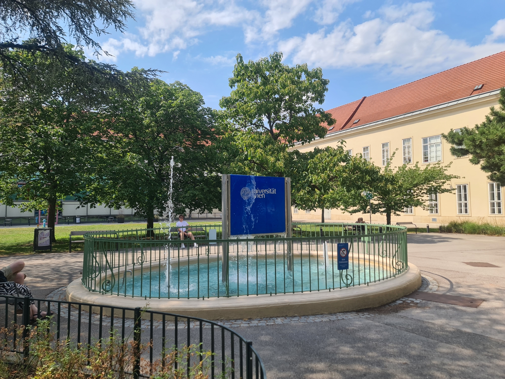 In Vienna, Austria, in front of a yellow building with a red tile roof, a circular fountain is active. The fountain is enclosed by a green metal fence with decorative scrollwork. A blue sign with white text that reads "Universität Wien" and a circular emblem is positioned behind the fountain. The sky is blue with scattered white clouds. Trees with green leaves are visible in the background. A young person wearing a white t-shirt and dark shorts is sitting on a bench to the right of the fountain, facing away from the camera. To the left of the fountain, a person is seated on a bench with their legs crossed, wearing a black and white patterned garment on their lower body. A black metal fence is in the foreground.