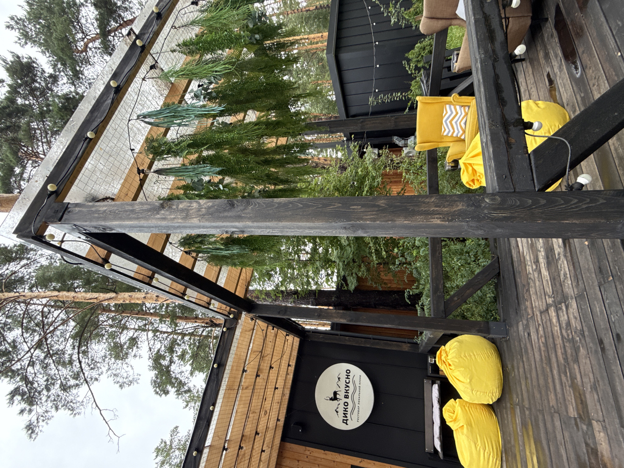 This image depicts an outdoor seating area of a restaurant named "Дико Вкусно" (Diko Vkrusno), which translates to "Wildly Delicious," located in Nizhnekayancha, Russia. The area is covered by a wooden and mesh roof supported by dark wooden beams, decorated with hanging artificial greenery and string lights.
On the wooden deck, there are two bright yellow beanbag chairs. To the left, a dark wooden building features a circular white sign with a logo of a deer and mountains, and text indicating it is a "РЕСТОБАР ЛОКАЛЬНОЙ КУХНИ" (Restobar of local cuisine). Further into the seating area, a bright yellow armchair with a gray and white chevron patterned cushion is visible, along with a brown armchair in the background. Behind the seating area, there is lush green foliage and trees, suggesting a natural, wooded environment. A dark, modern-looking structure is partially visible on the right side of the image. The wooden deck appears damp, possibly from recent rain. There are no people visible in the image.