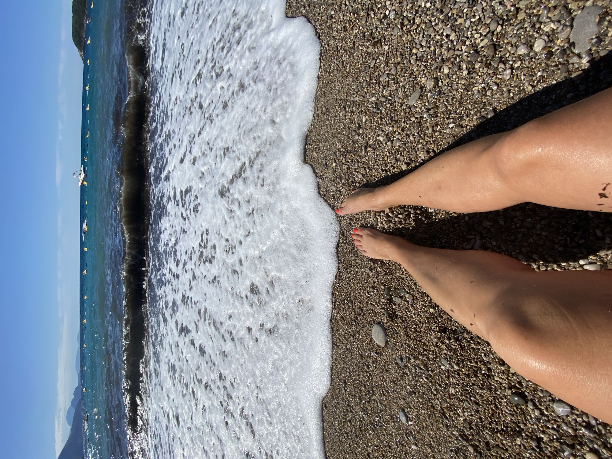 A first-person perspective shot shows a person's legs and feet resting on a pebble beach, with the ocean and sky in the background. The water is a deep blue and turquoise, with white foam from a breaking wave washing up on the shore. There are yellow buoys visible in the water, and a small boat in the distance. Mountains are visible in the far background. The sky is clear and blue. The scene is located in Prijevor, Montenegro.