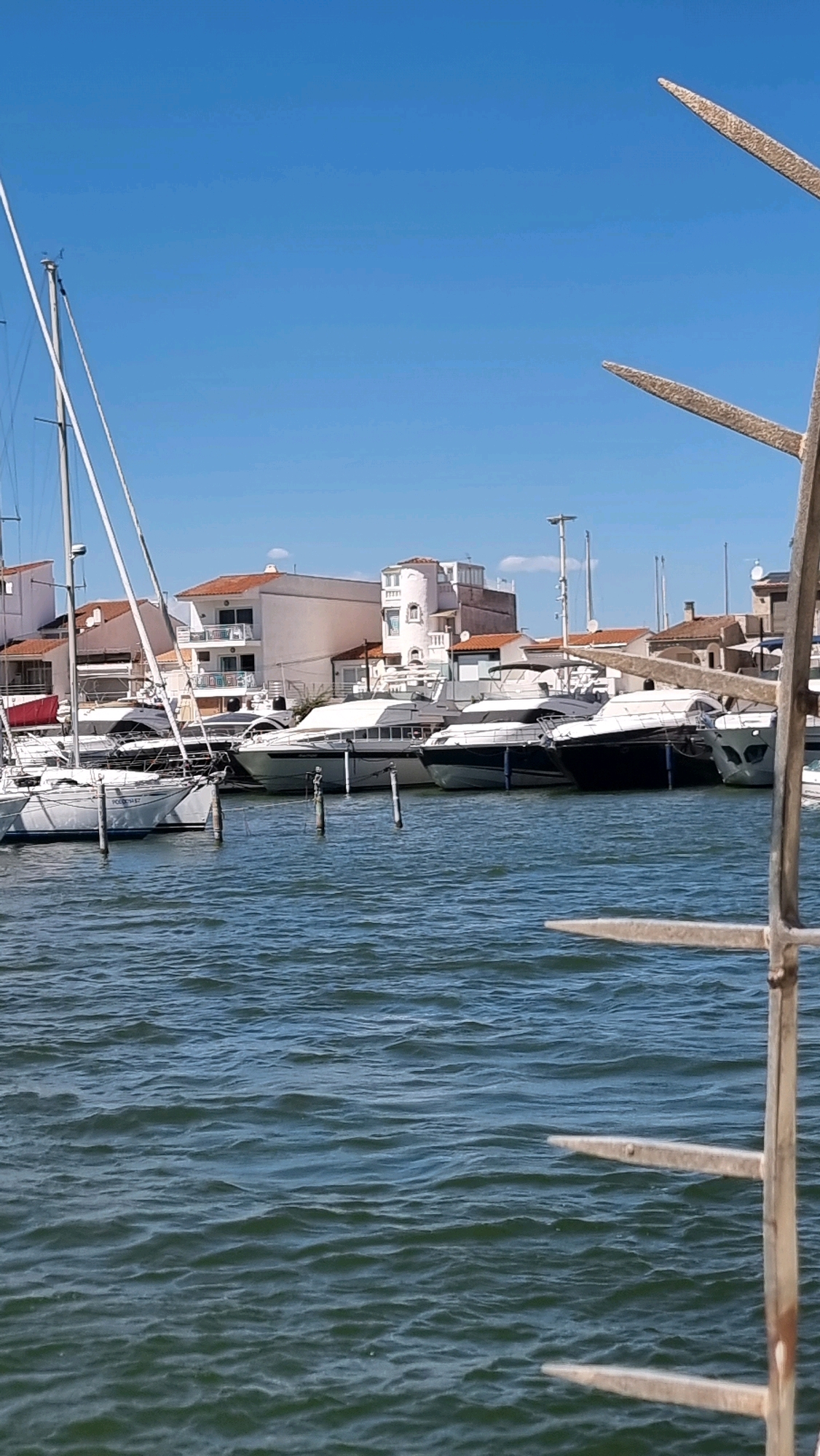 The image shows a marina in Castelló d'Empúries, Spain, under a clear blue sky. Several white yachts and sailboats are moored in the calm, blue water. Buildings with white walls and orange-tiled roofs line the waterfront. In the foreground, a portion of a metal sculpture with pointed prongs is visible on the right side of the frame, partially obscuring the view of the boats and buildings. The water is rippled, indicating a gentle breeze. There are no people visible in the image.