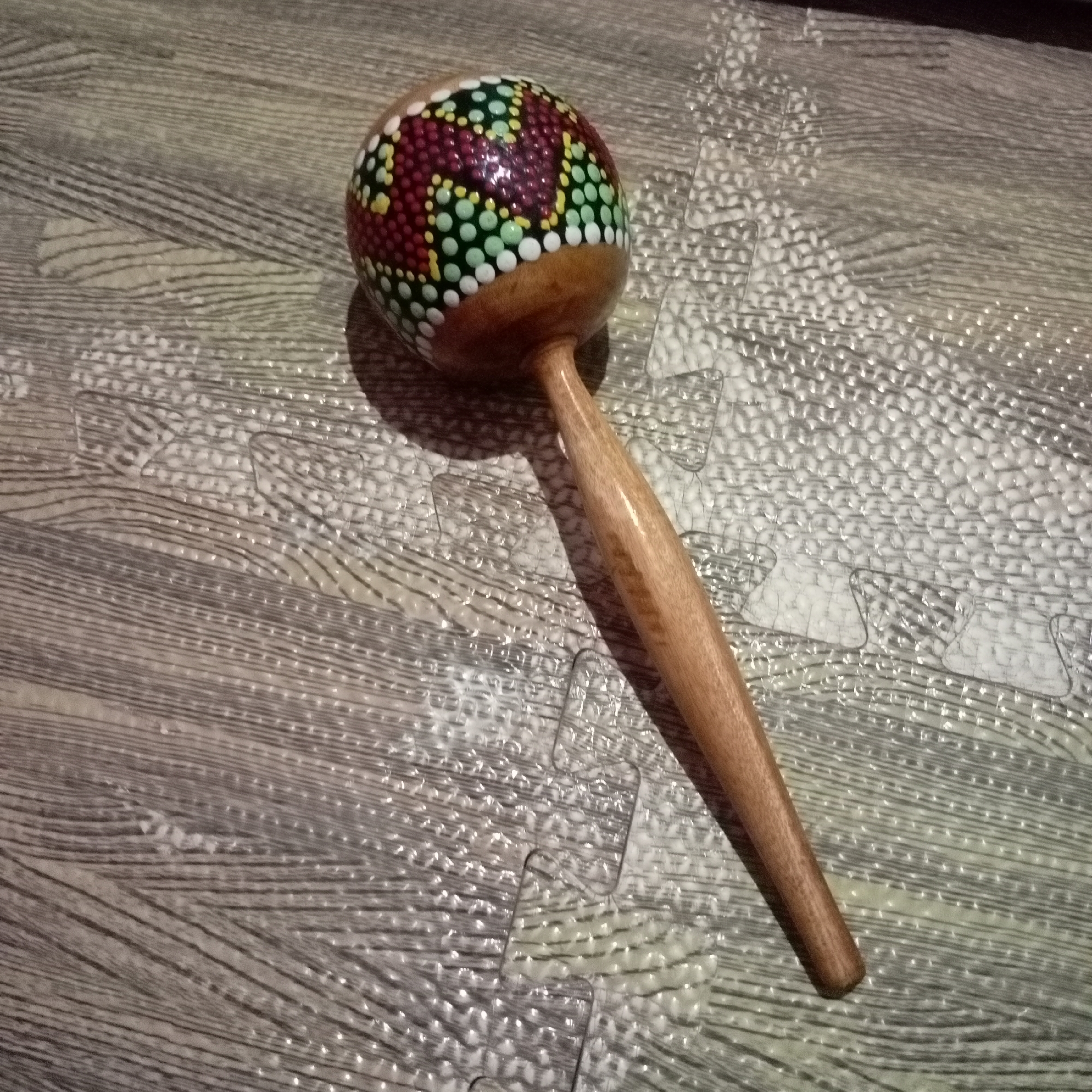 A single maraca lies on a grey, textured surface. The maraca has a round, wooden head decorated with colorful dots in red, yellow, green, and white, arranged in a patterned design. It has a smooth, brown wooden handle that extends downwards from the head. The surface it rests on appears to be a mat with a wood-grain pattern and an interlocking, puzzle-like design visible in some areas, possibly a floor mat or padded surface. The lighting casts shadows on the surface, indicating a light source from above and to the left of the maraca. There are no people or animals present in the image, and no specific city or location is identifiable from the visual information provided.