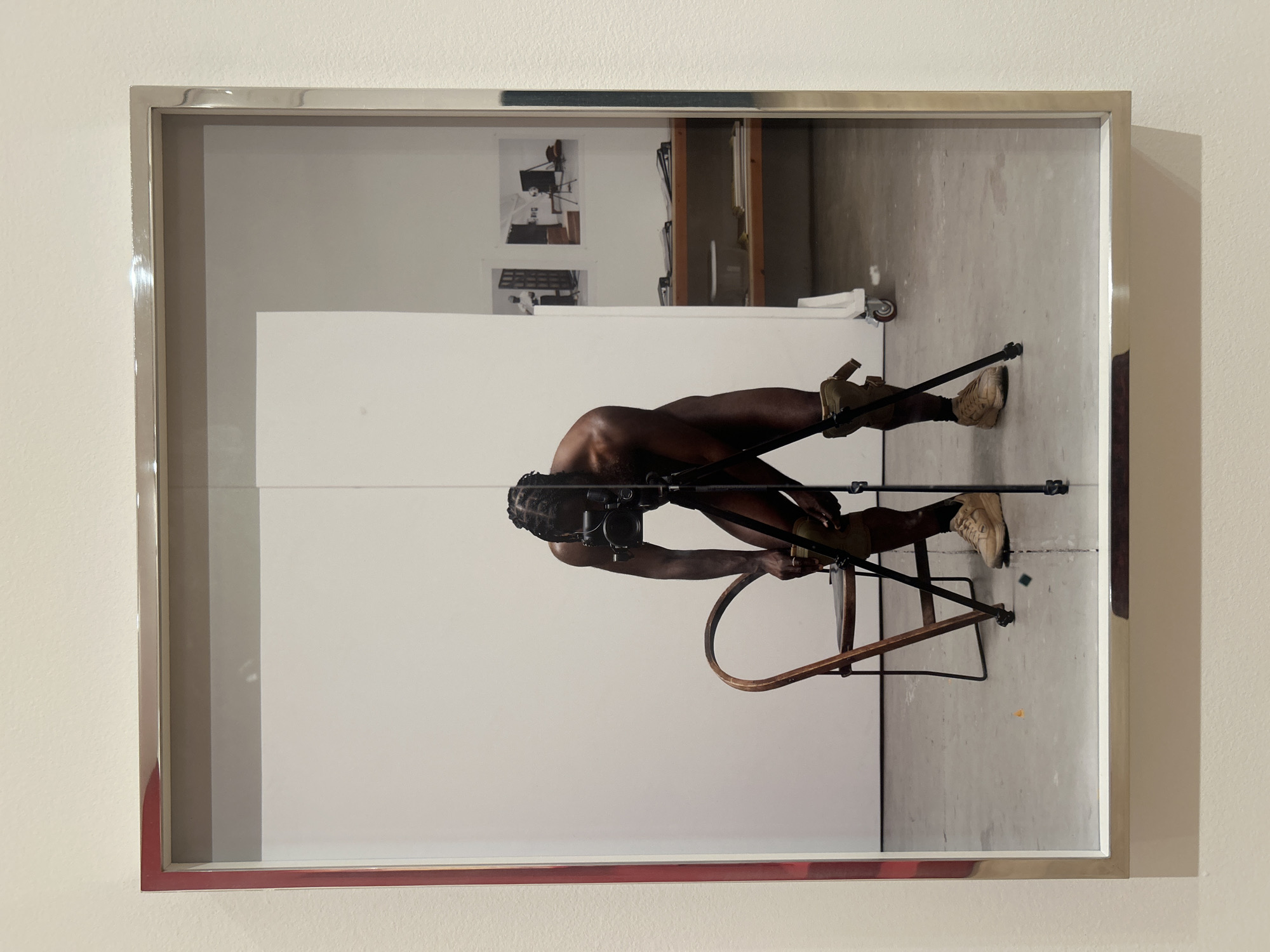 The image depicts a photograph in a silver frame hanging on a light beige or white wall. The photograph appears to show a nude person with dark skin, standing on an upside-down chair while holding a camera. The background consists of a white partition with two photographs hanging above it. Beyond the partition, there's a glimpse of a shelving unit. The scene seems to be set in a studio-like environment with concrete floors. The reflection in the glass obscures details. The location is unknown, but is suspected to be in Los Angeles, United States of America.