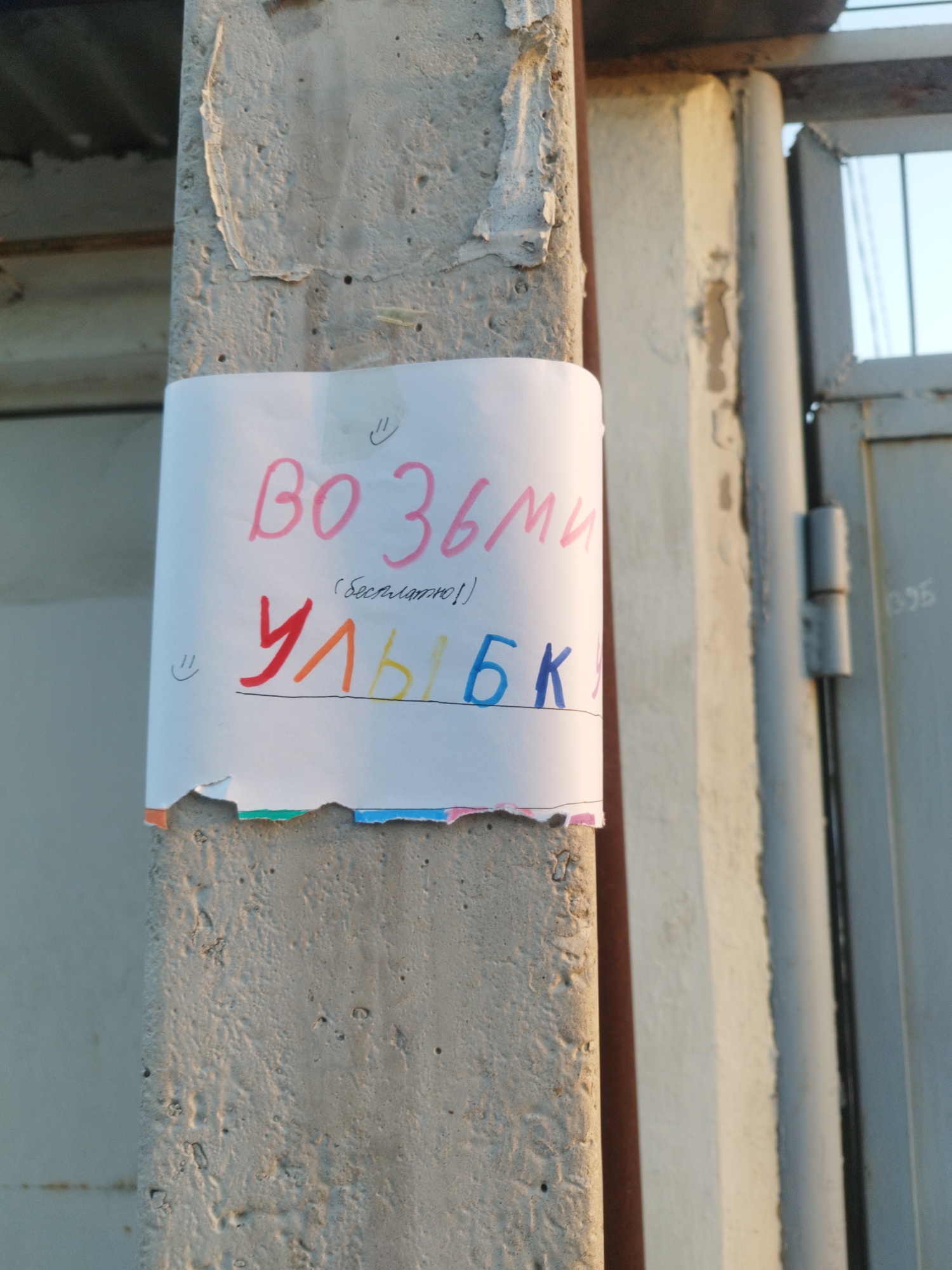 A hand-drawn sign on white paper is taped to a concrete pillar in an outdoor urban setting. The sign features handwritten text in pink and blue ink. The larger, top line of text, written in pink, appears to say "ВО ЗЬМИ" which translates to "TAKE". Below this, in smaller black text, are the words "(бесплатно!)", meaning "(free!)". The bottom line of text, written in a mix of red, orange, yellow, and blue, spells out "УЛЫБКИ", meaning "SMILES". Two small smiley faces are also drawn on the paper, one above the first line of text and one to the left of the second line. The bottom edge of the sign is torn and shows colored paper fragments. The concrete pillar is rough and textured, with some peeling paint on its surface. In the background, a portion of a grey metal gate and a vertical metal pipe are visible, suggesting the location is an entrance or boundary in a public space. The lighting indicates it is daytime, possibly late afternoon due to the warm tones. The sign is positioned at a height accessible to pedestrians. Based on the provided metadata, this scene is located in Shymkent, Kazakhstan.