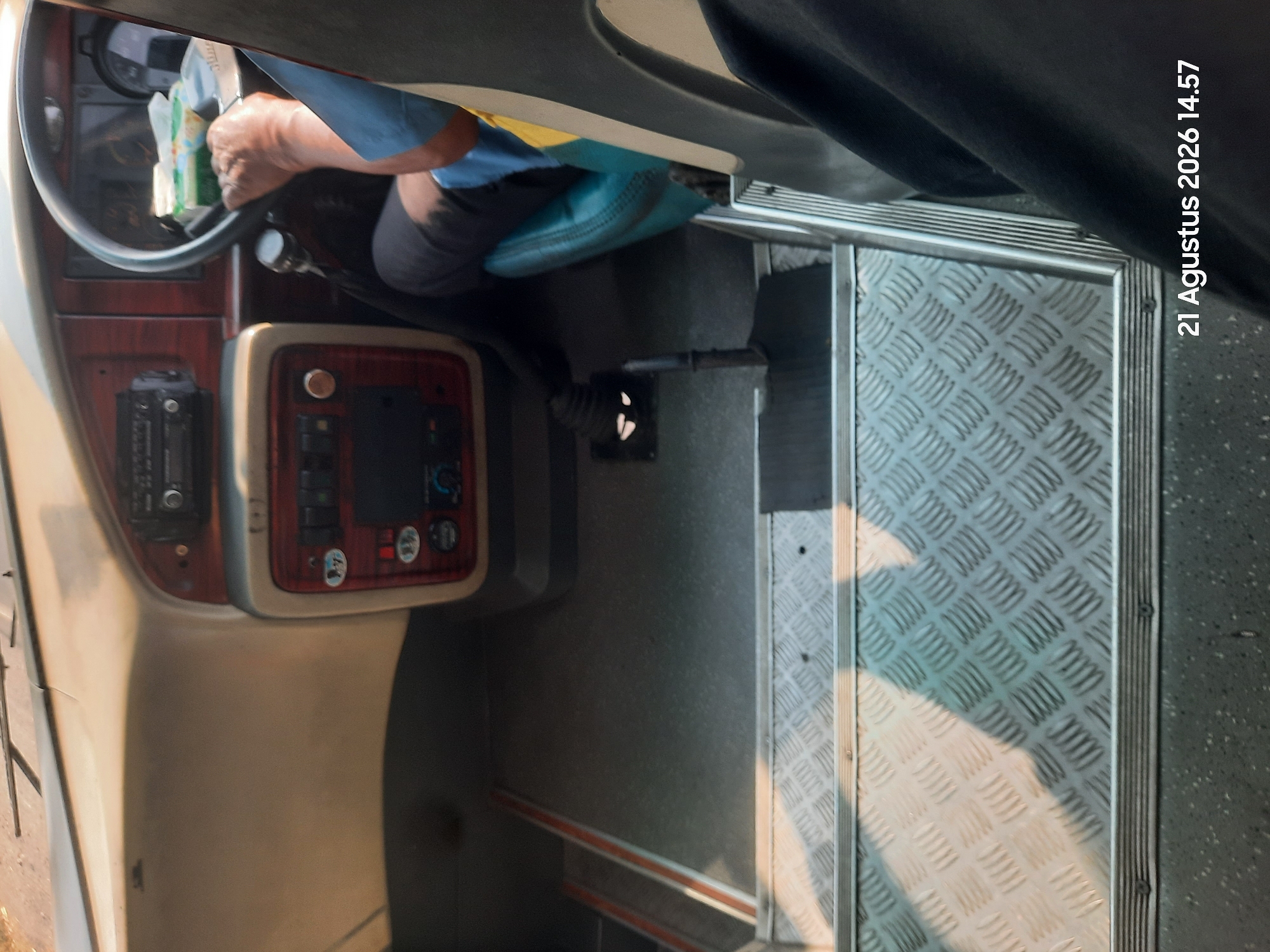 The image shows the interior of a bus, viewed from the passenger area looking towards the driver's seat. The driver, a person with dark skin and wearing a blue shirt, is visible at the steering wheel, holding it with their right hand. A box of tissues is placed on the dashboard near the steering wheel. The bus has a dashboard with various controls, including what appears to be a radio and climate control buttons. The floor of the bus is visible, with metal tread plates forming the steps leading up to the driver's area. Sunlight casts shadows across the floor. A date and time stamp, "21 Agustus 2026 14.57", is present in the lower right corner of the image. The location is identified as Sukokerto, Indonesia.