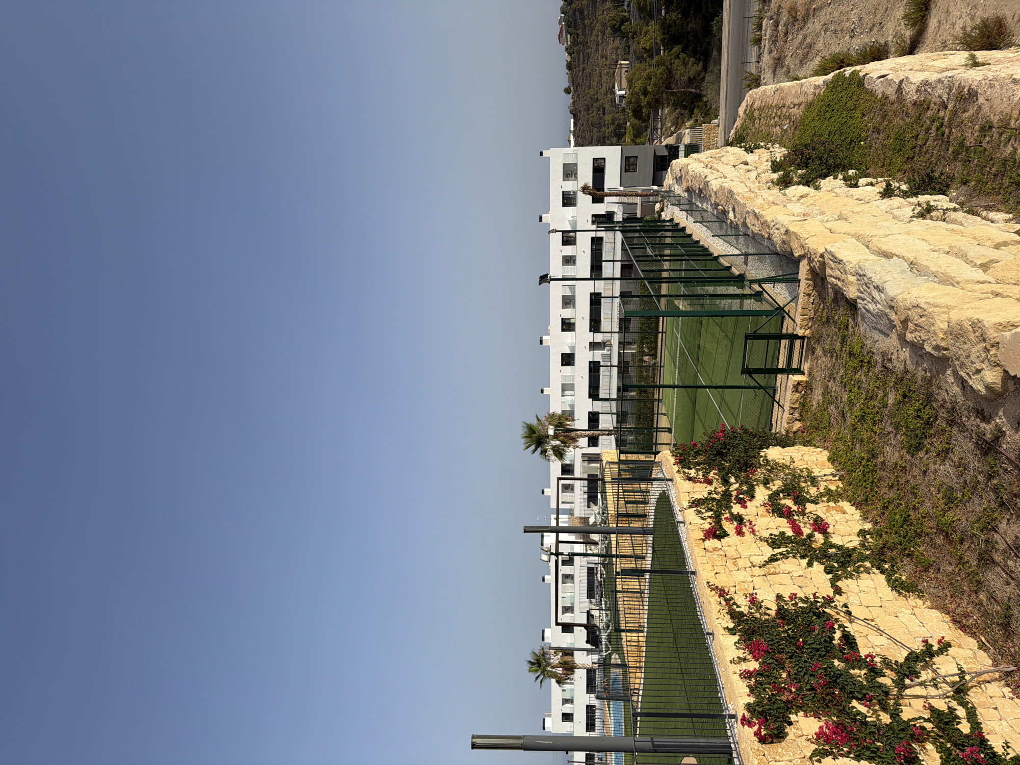This is a medium-shot, eye-level, horizontal photograph taken outdoors in Finestrat, Spain, featuring a modern apartment complex and recreational facilities. The scene is dominated by a clear, pale blue sky that occupies the upper half of the frame. Below the sky, a long, white, multi-story apartment building stretches across the middle ground. This building features numerous windows with dark frames and small balconies. In front of the building, there is a swimming pool with blue water on the left side, and two fenced green sports courts, likely for paddle tennis, on the right side.
The foreground is characterized by a tiered landscape. A low, rough-hewn stone wall, with vines bearing bright pink flowers growing on it, forms the base of the foreground. This wall is made of large, unevenly shaped blocks of light-colored stone. Beyond the stone wall, there are patches of dry, sparse vegetation and soil, suggesting a semi-arid environment. The sports courts are enclosed by dark green metal fencing. Two tall, grey lampposts stand on either side of the scene, in front of the apartment building. In the background, a hilly, vegetated landscape with scattered houses and trees can be seen, and further in the distance, a hazy horizon suggests the sea or a large body of water. The lighting indicates it is daytime, likely late morning or afternoon, with the sun casting subtle shadows. There are no people visible in the image.