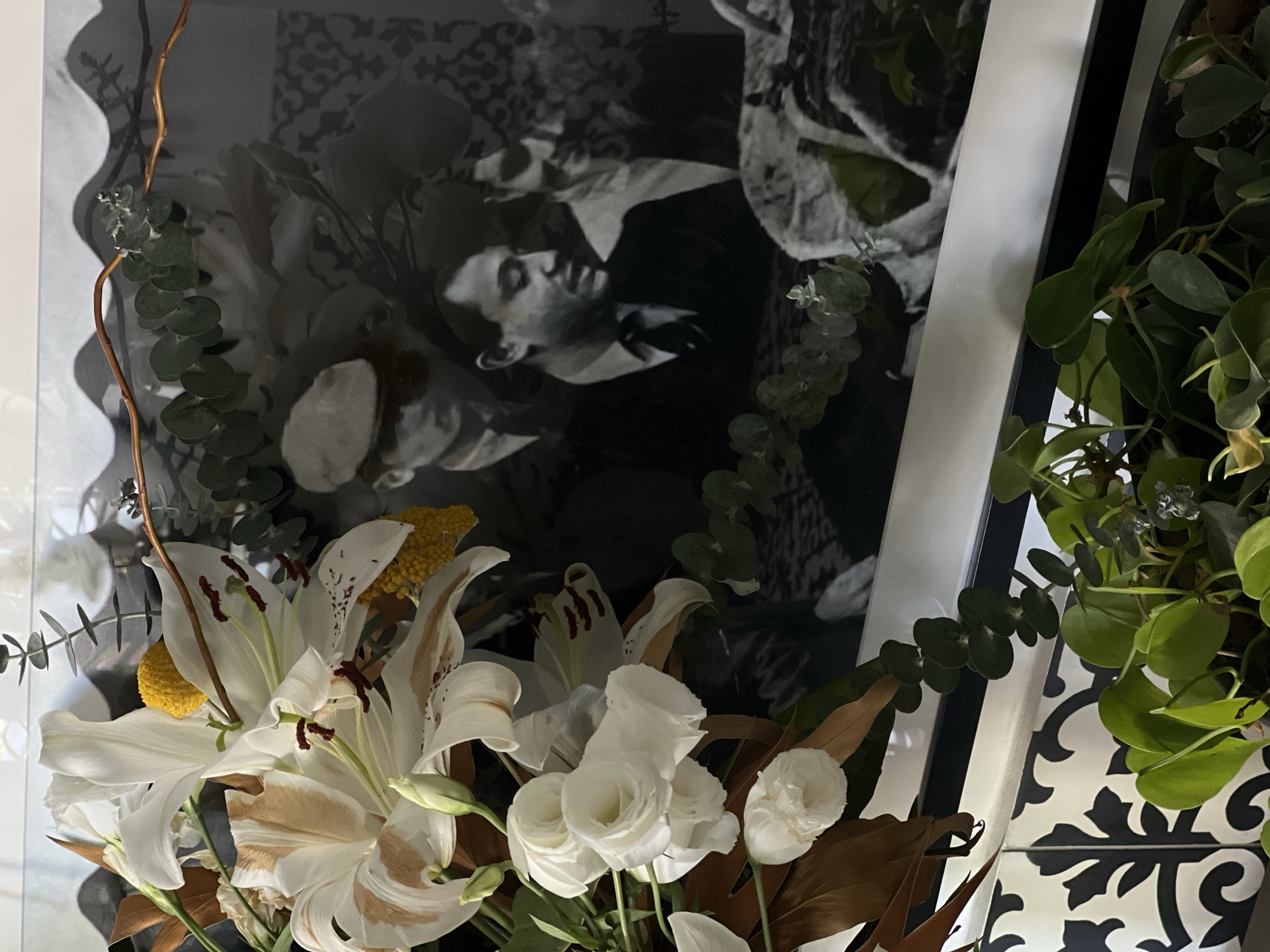 The image shows a still life arrangement with white lilies and roses in the foreground. Behind the flowers is a black and white framed photograph of a man in a suit. The background is partially obscured, but reveals a white and black patterned wall or tile. There is no discernible information in the image that indicates this scene is in Tel-Aviv, Israel.