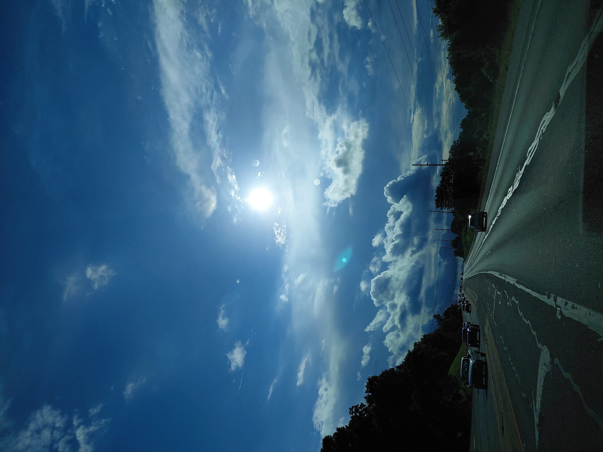 The image shows a view from a vehicle driving on a highway in Versailles, United States. The sky is a deep blue with dramatic clouds, and the sun is visible, creating lens flare. Trees line the left side of the highway, and there are power lines visible in the distance on the right. Several cars are also on the highway, driving in the same direction. The road surface shows some wear and tear with cracks and patches. The overall impression is of a late afternoon drive under a dynamic sky.