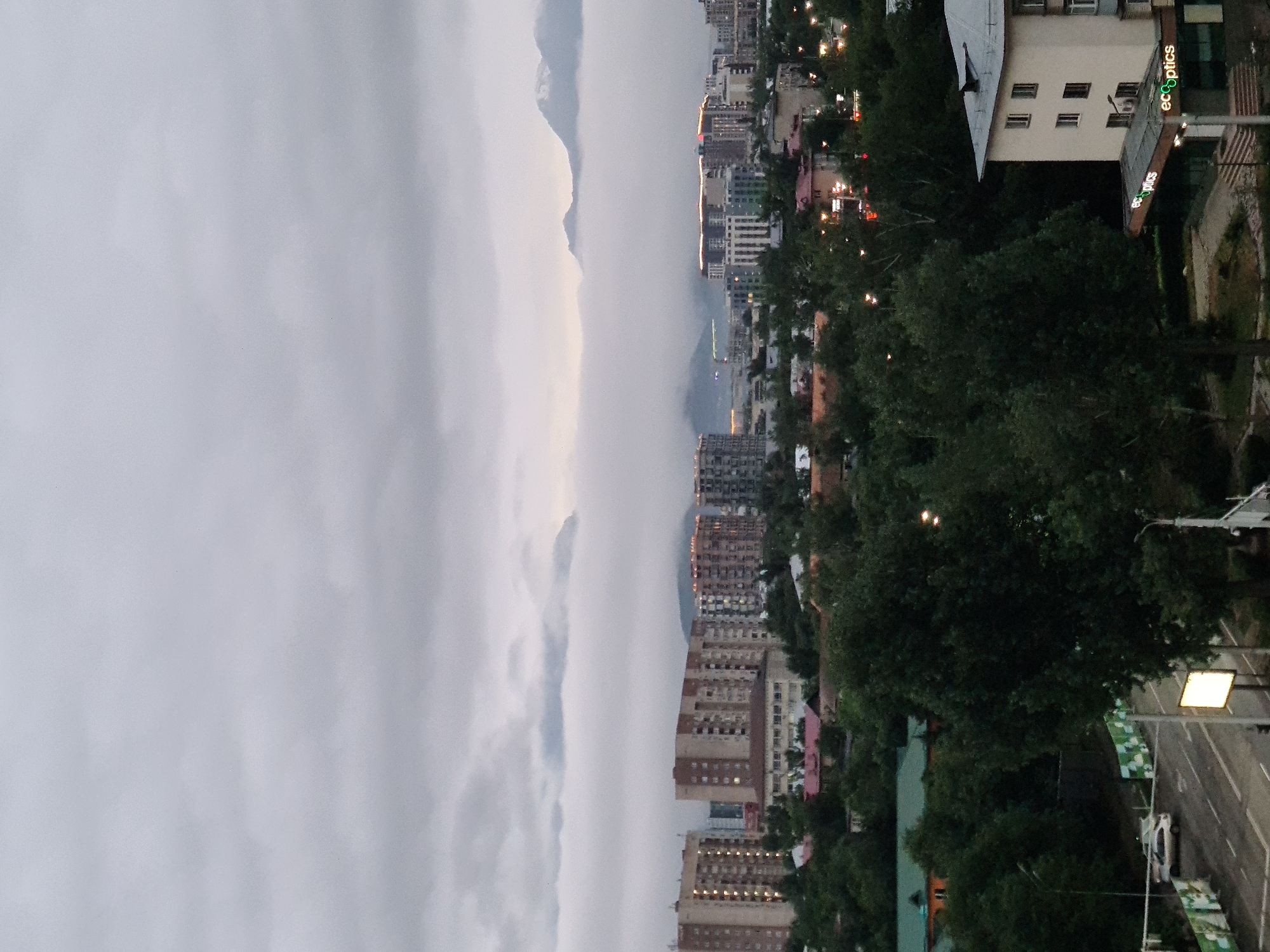 The image captures a panoramic view of a city with mountains in the background, under a cloudy sky. The foreground features lush green trees, a road with a few vehicles, and several buildings. In the mid-ground, a cluster of apartment buildings and other structures are visible, some illuminated with lights. The background is dominated by mountains partially obscured by fog or low-lying clouds, with the peaks of the mountains peeking through. The overall atmosphere is serene and slightly overcast, suggesting it might be early morning or late evening. The architecture of the buildings and the presence of the mountains strongly suggest the location is Almaty, Kazakhstan, a city known for its proximity to the Tian Shan mountain range. Specifically, the cluster of modern apartment buildings with illuminated facades and the distinct shape of the mountains hint at the area near the city center or its outskirts adjacent to the mountains. There are no people clearly visible in the frame. The scene appears to be a typical urban landscape in Almaty, characterized by a blend of modern development and natural mountain scenery.