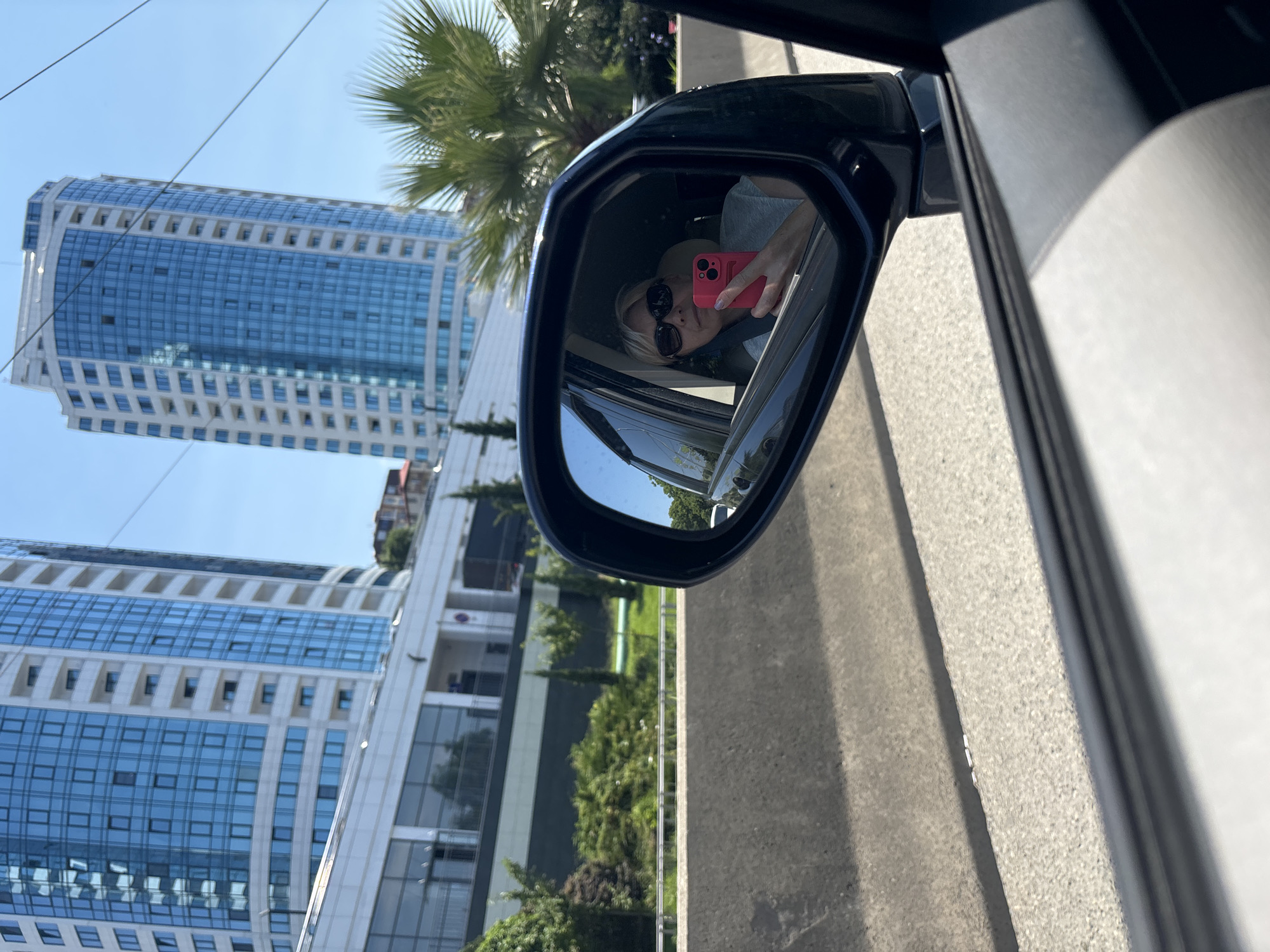 The image captures a sunny day in Sochi, Russia, likely near a modern coastal development. In the foreground, a car's side mirror reflects a blonde woman with sunglasses taking a selfie with a pink smartphone. She is wearing a light-colored top. Behind her, the passenger side window of the car is visible. The background reveals two prominent, tall, modern buildings with blue glass facades, characteristic of recent construction in urban centers. Palm trees and lush green foliage, including cypress trees, are interspersed between the buildings and along the roadside, suggesting a warm climate. The road appears to be a multi-lane highway, separated by a concrete barrier. Overhead power lines are faintly visible against the clear blue sky.