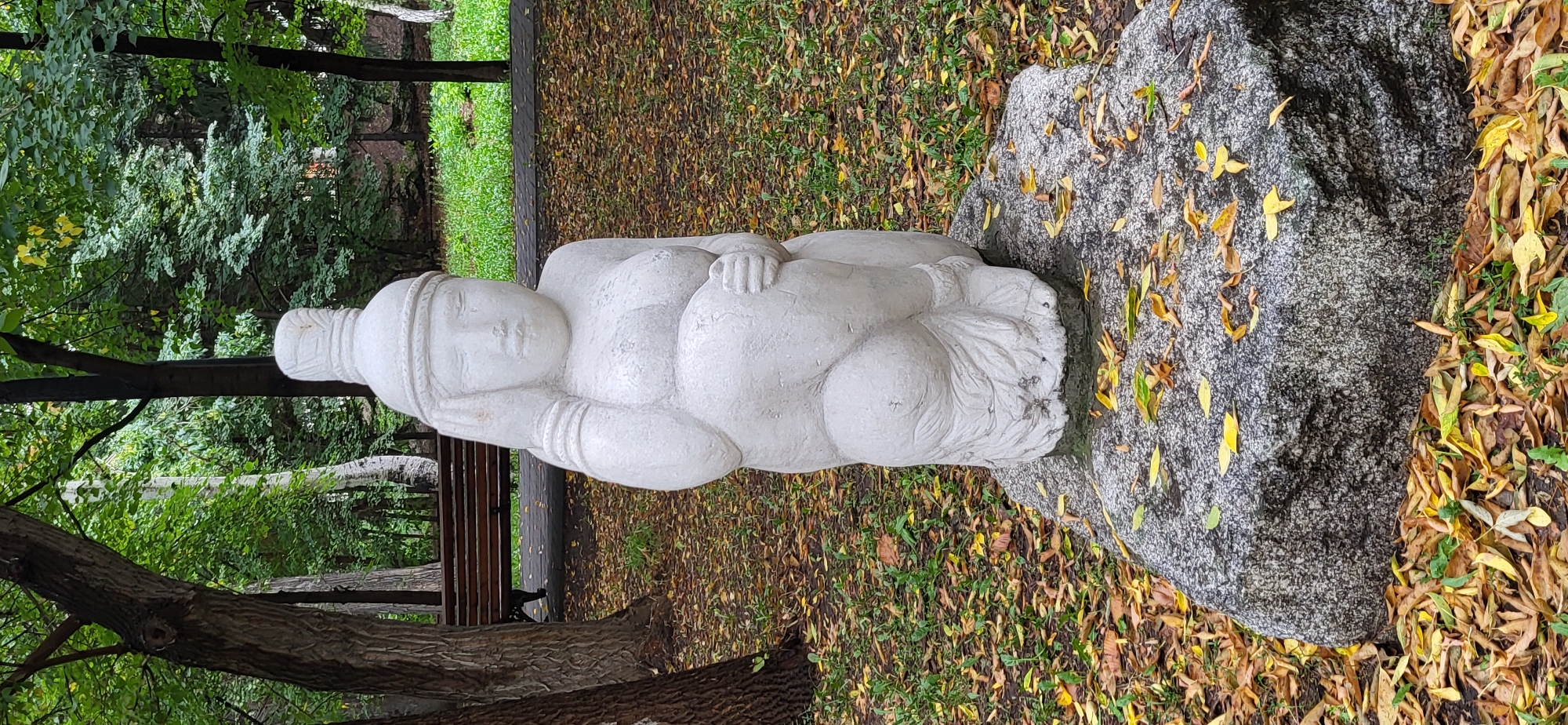 A stone sculpture of a seated female figure, possibly depicting a mother or pregnant woman, is positioned on a large, textured granite boulder in a park setting. The sculpture is light-colored and appears weathered. The figure has a rounded body, is holding its belly with one hand, and has its other hand raised to its head, touching its temple or ear. The figure wears a headdress and armbands. Surrounding the boulder are fallen autumn leaves in shades of yellow, brown, and orange, scattered across a green, grassy ground cover. In the background, trees with green and yellow foliage form a dense canopy, with a wooden park bench visible behind the sculpture. The overall scene suggests a peaceful, outdoor environment. This sculpture is located in the city of Oskemen, Kazakhstan, specifically in the park on Kirov Street, near the regional akimat (administration building).