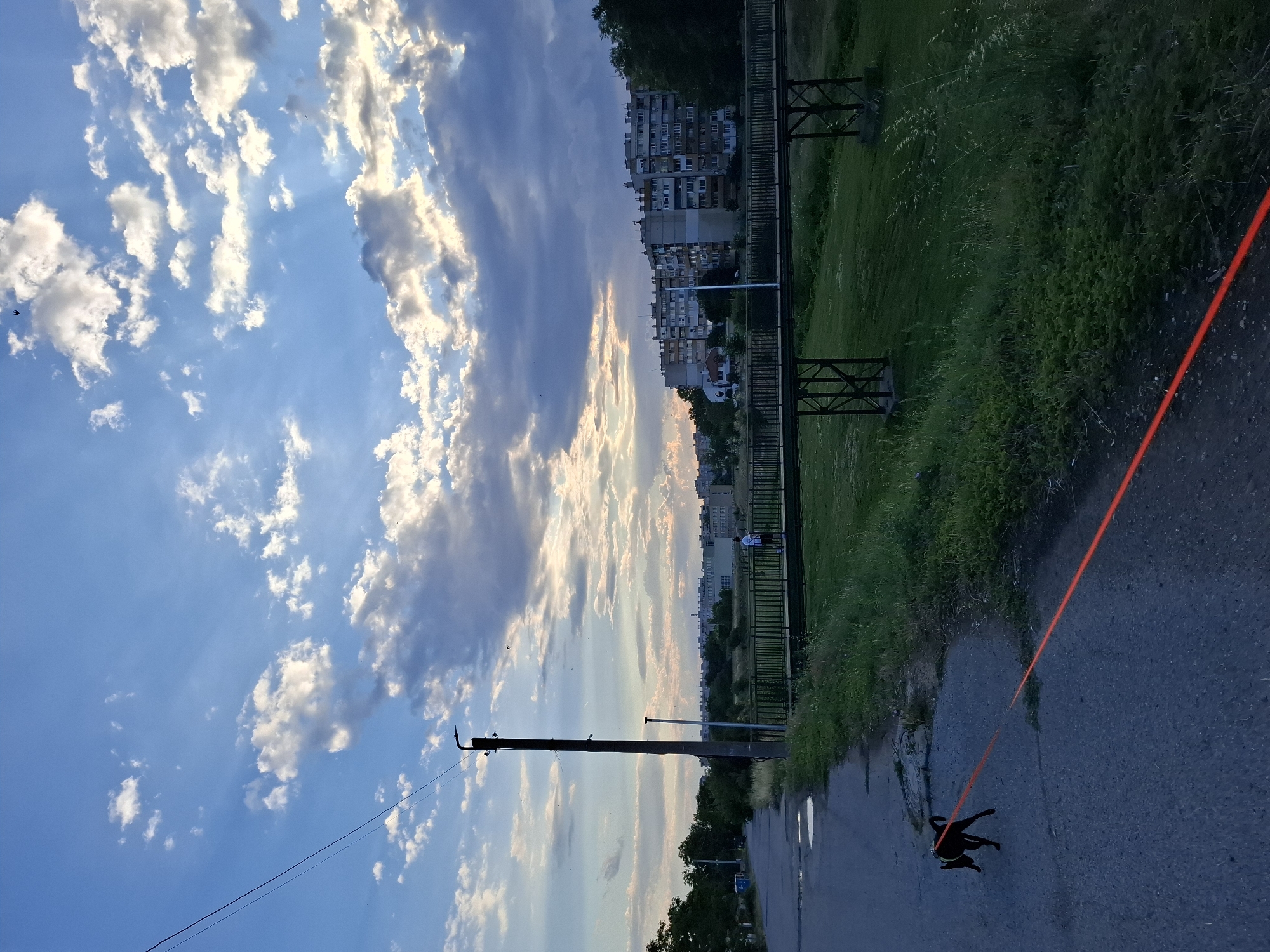 An evening scene captures a street in Yambol, Bulgaria, as a small, dark dog with a red leash walks on a paved path. The dog is silhouetted against the asphalt, and the leash stretches diagonally across the foreground. In the background, a pedestrian bridge spans a grassy area, with a person walking on it. Beyond the bridge, apartment buildings line the horizon under a sky filled with dramatic clouds, illuminated by the setting sun. A utility pole stands to the left of the dog, casting a shadow on the road. The environment appears to be a mix of urban development and green spaces.