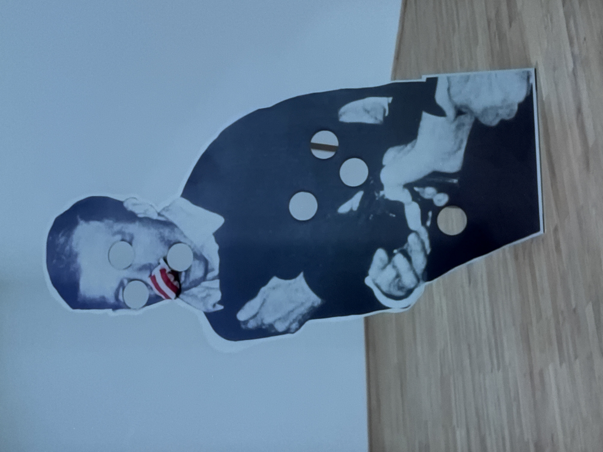 The image displays a life-sized, monochromatic cutout of a male figure, standing upright on a light wooden floor against a plain, light-toned wall. The figure, dressed in a dark, collared long-sleeved garment, features several circular cutouts: two on the face obscuring the eyes, one obscuring the mouth, and additional ones on the torso. A red and white striped, conical object is inserted into the facial cutout at the approximate nose position. The figure's left hand is raised towards the chest. The right hand is situated lower, adjacent to a smaller, secondary image on the cutout's lower right quadrant, which depicts the lower body and legs of another person. This scene is situated within an indoor exhibition or gallery environment in Riga, Latvia.