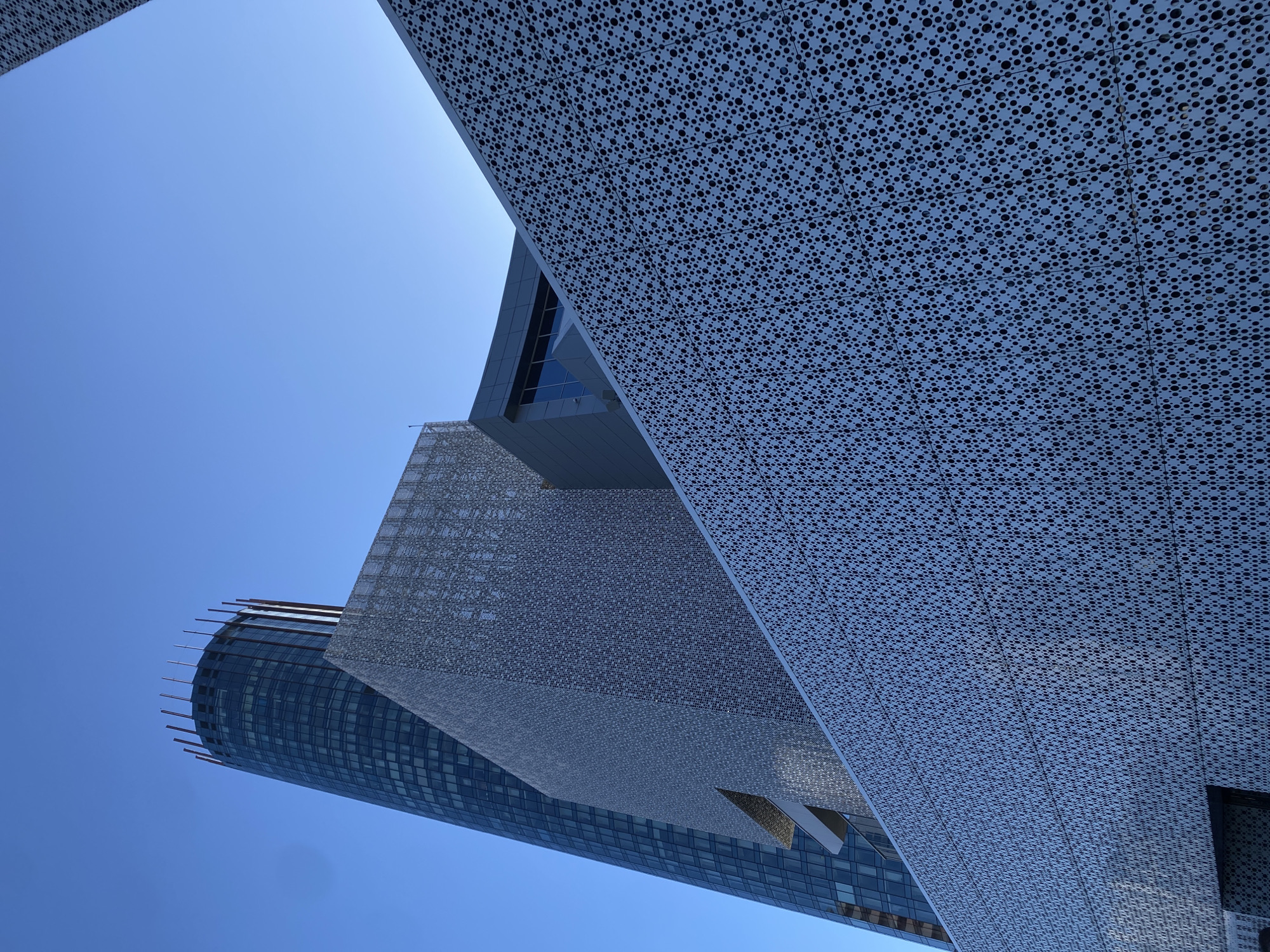 The image shows a low-angle view of several modern buildings in Yekaterinburg, Russia, set against a clear, blue sky. Dominating the foreground are structures featuring a perforated screen-like facade. These facades are detailed with a consistent, repeating geometric pattern of small holes. Behind these, a cylindrical skyscraper with a glass curtain wall rises. Horizontal elements are visible on the roof of the skyscraper, likely acting as architectural details. The structures appear to be juxtaposed with sharp angles and intersecting lines, creating a complex interplay of shapes and textures. The overall impression is one of modern architectural design in an urban setting.
