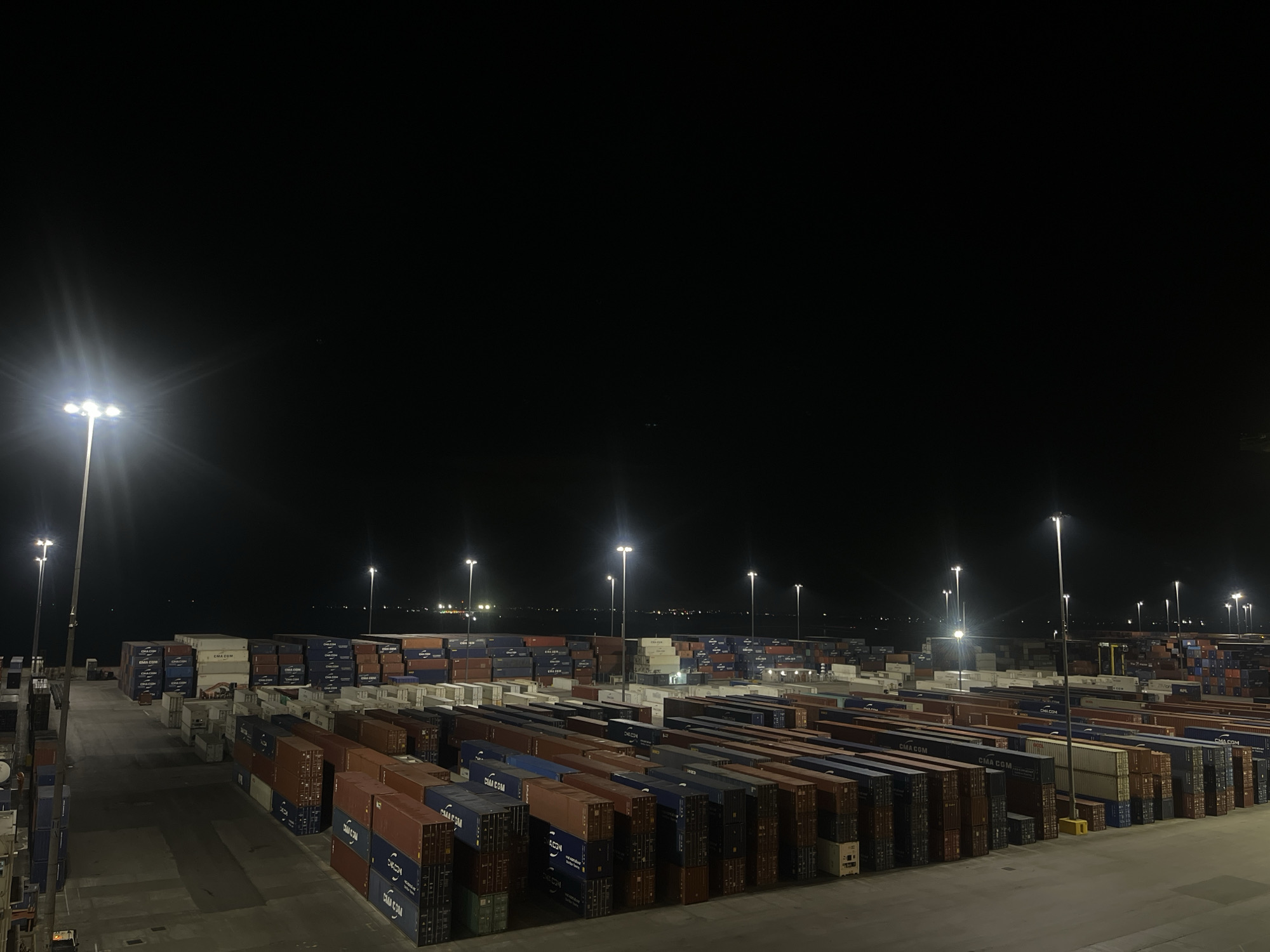 The image shows a container terminal at night, presumably a port or shipping yard. The scene is illuminated by tall, bright floodlights which cast light on the containers. Rows of shipping containers fill the foreground and midground, stacked in an organized manner. The containers are of various colors, including blue, red, white, and grey. The ground is paved and shows the wear of heavy equipment and movement. The sky is black. The location is unknown, but it is likely an industrial area near water based on the presence of containers.