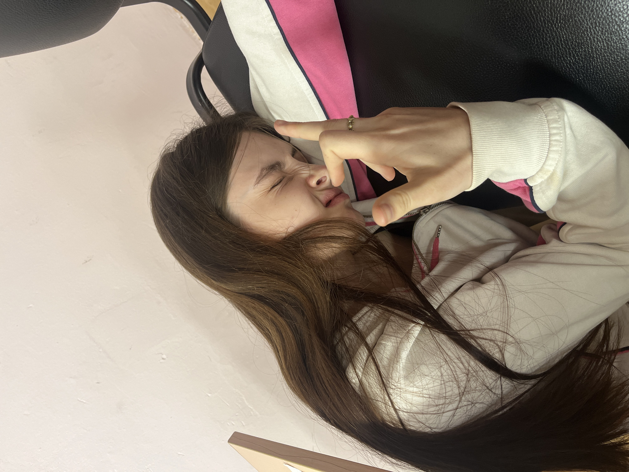 A person is pictured lying on a chair. The person has long brown hair, pale skin, and is wearing a white jacket with pink and navy trim. They are making a gesture with their hand near their face, and they are wearing a ring on their finger. The background includes a black chair, a white wall, and a hint of what appears to be a desk or table. There is no evidence to determine the location in the city of Saint Petersburg, Russia.