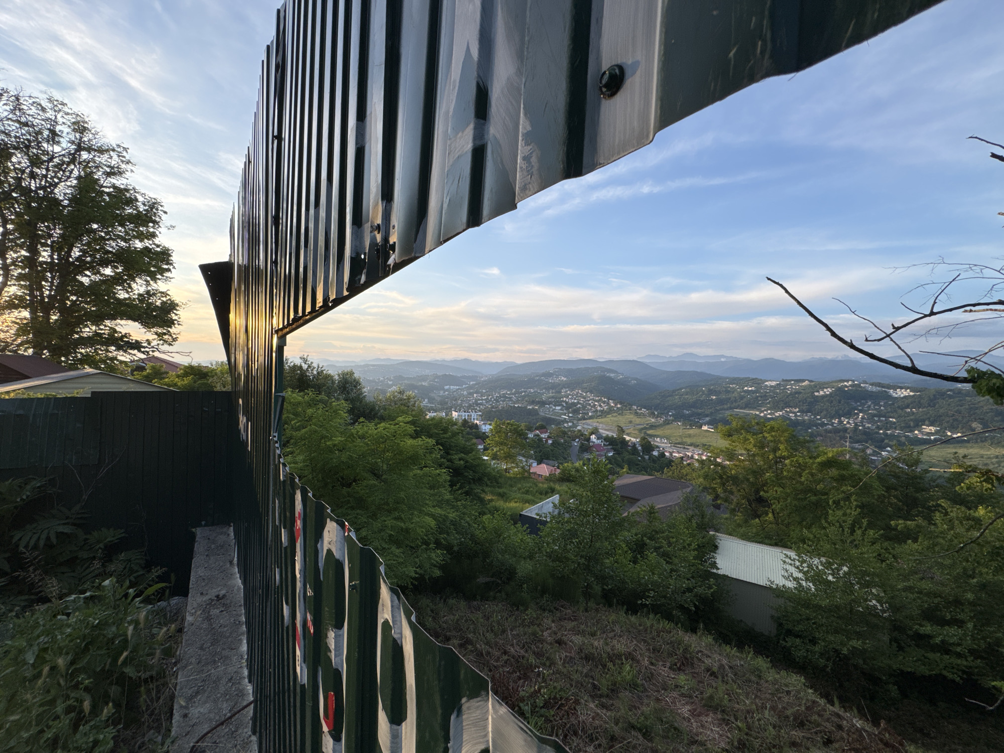 The image captures a scenic overlook of the city of Sochi, Russia, from a vantage point framed by a corrugated metal structure and fence. The sun is setting, casting a warm glow on the sky and distant mountains.
In the foreground, a dark green corrugated metal fence with red and white painted markings curves along the edge of the frame, partially obscuring the view. To the left of the fence, a concrete retaining wall is visible, covered with overgrown vegetation.
Beyond the fence, the landscape unfolds with a dense green forest covering the hillside. A few houses with brown and white roofs are nestled amongst the trees. The city of Sochi stretches out in the valley below, with buildings and roads winding through the hilly terrain. The distant mountains are shrouded in a soft haze, with the setting sun creating a warm, orange glow on the horizon.
The sky is a gradient of blue, with wispy white clouds scattered across it. A single bare tree branch extends into the frame from the top right corner, adding a touch of nature to the urban landscape. The overall impression is one of peaceful coexistence between the natural environment and urban development, with the sunset adding a dramatic and beautiful backdrop.