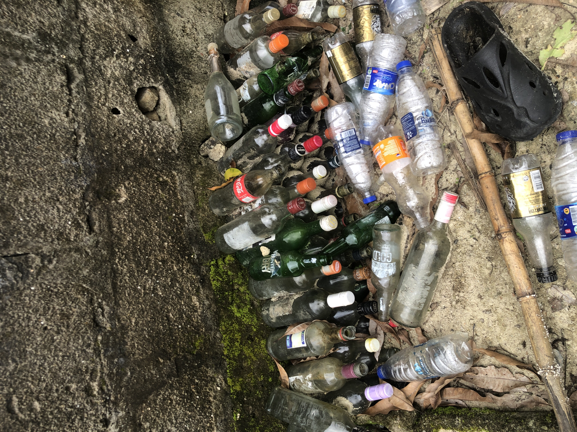 A pile of discarded glass and plastic bottles, along with a black rubber sandal, lie on sandy ground against a rough concrete wall. The wall has patches of moss and some small holes. The bottles are of various shapes, sizes, and colors, including clear, green, and brown glass, and clear plastic. Some bottles have labels and caps, while others are empty. The scene appears to be an outdoor location, possibly a roadside or a less-trafficked area, suggesting a waste disposal or littering issue. The lighting suggests it is daytime. The image does not depict any people or animals. The location is identified as Ado, Nigeria.