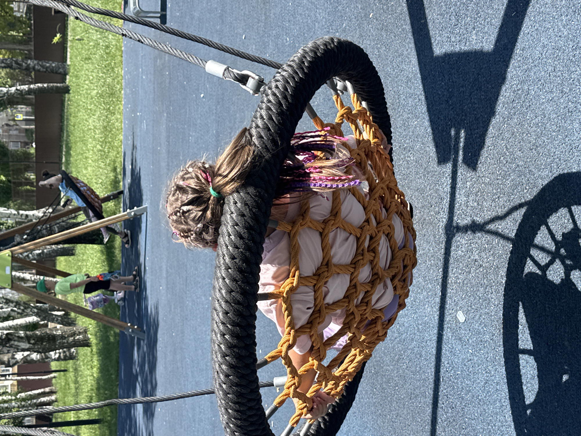 In Mikhaylovsk, Russia, a young girl with braided hair, adorned with colorful streaks of pink and purple, is enjoying a sunny day at a playground. She is seated in a circular net swing, its thick black rope frame contrasting with the woven tan netting that cradles her. The girl wears a light pink top and purple shorts. The swing is suspended by thick cables, indicating it's part of a larger play structure.
The playground has a soft, blue rubberized surface, typical of modern play areas, providing a safe landing zone. To the left of the swing, a section of bright green grass is visible, bordered by a dark brown fence. Several birch trees with their distinctive white bark and green foliage are scattered throughout the background, suggesting a park or green space. In the distance, a hint of buildings can be seen.
A shadow of the swing's structure, with a distinct circular and spoke-like pattern, is cast on the blue ground, indicating it's a bright, sunny day, likely mid-morning or afternoon. Another child, wearing a bright green shirt and a green cap, is visible in the background, standing near a wooden play structure and holding a toy. The overall atmosphere appears cheerful and relaxed, with children enjoying outdoor recreational activities. No visible text is present in the image.