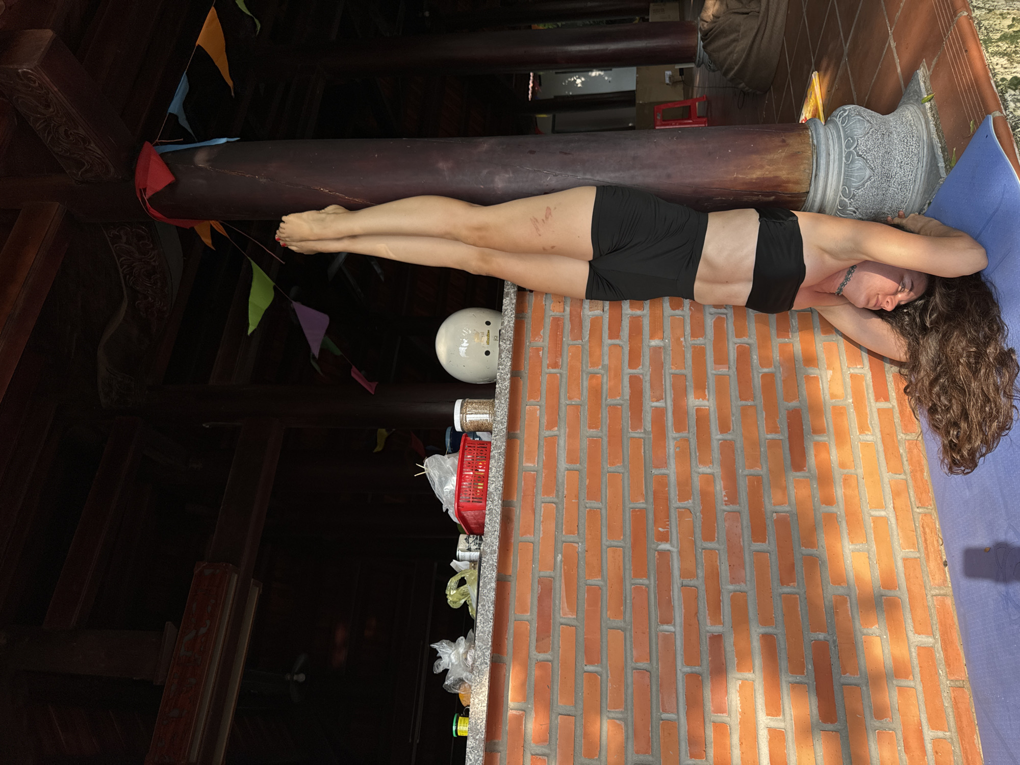 An adult individual with long, curly dark hair performs a supported inversion pose (headstand or forearm stand) on a blue yoga mat in Mũi Né, Vietnam. The person is dressed in a black sports bra and black shorts, with their legs extended vertically, resting against a dark wooden pillar. Scratches or marks are visible on the upper left thigh. The pose is executed on an outdoor tiled or paved surface, adjacent to a red brick wall. Above the brick wall is a light-colored counter holding a white helmet, various plastic bags, a red basket, and several jars. Colorful triangular bunting is strung between the wooden pillars, which feature ornate carved stone bases and decorative elements near the ceiling. A darker wooden ceiling structure with beams is visible overhead. Strong sunlight illuminates the brick wall and floor from the left, casting distinct shadows. In the background, a red stool and a brown bean bag are visible.