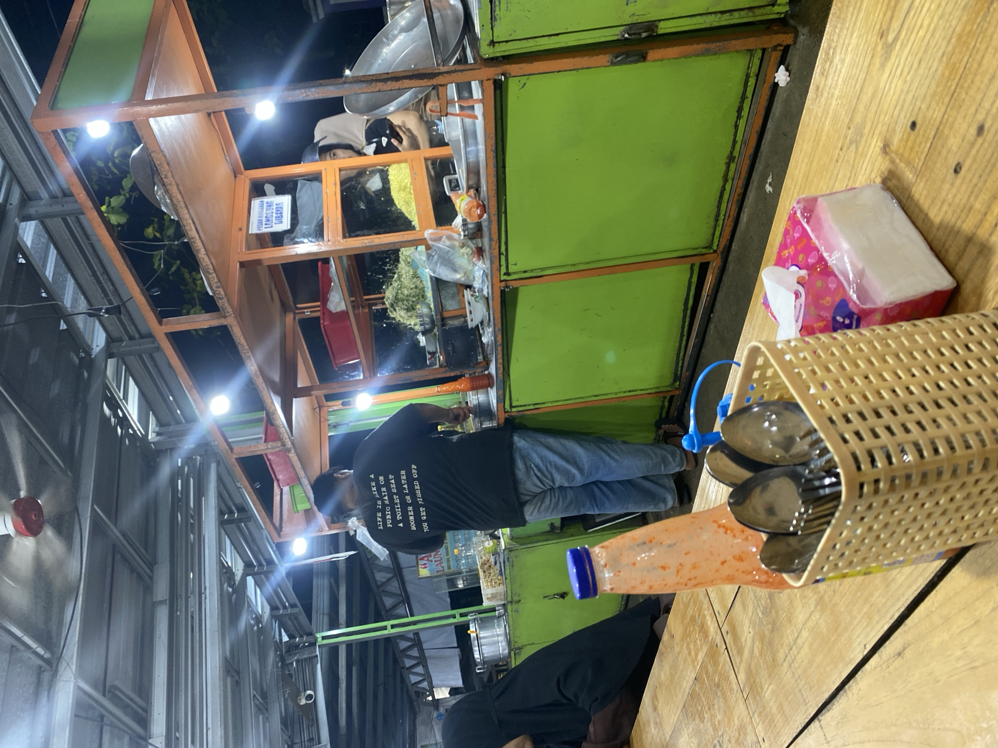 The image depicts an outdoor food stall at night in Banjarbaru, Indonesia. A man wearing a black t-shirt with text and blue jeans stands behind a green and orange food cart, actively preparing food. To the left, a person wearing a black hijab is seated, partially visible. On the wooden table in the foreground, there is a basket of cutlery, a bottle with an orange cap, and a pack of facial tissues. The food cart is illuminated by overhead lights, revealing various food ingredients and cooking utensils. The overall scene suggests a bustling evening street food environment.