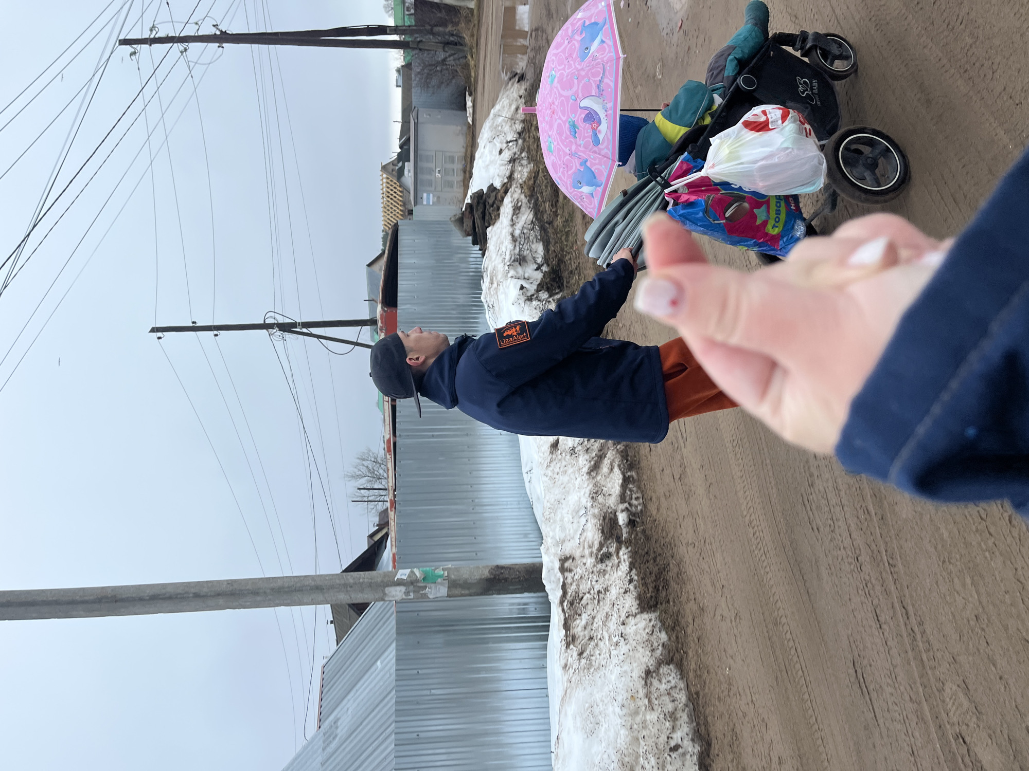 The image captures a scene in Chaikovsky, Russia. A man wearing a navy blue jacket with a "LizaAlert" patch on the sleeve, an orange undershirt, and a dark cap, stands on a muddy road next to a snowbank. He is holding an umbrella with a pink dolphin design. Beside him is a black baby stroller filled with items covered in a green blanket, with a white plastic shopping bag on top of it and a patterned blue shopping bag in front of the bag. The road is covered in tire tracks. A partial view of a person’s hand wearing a matching blue jacket is in the lower right corner of the photo.
The background includes a corrugated metal fence and various buildings under an overcast sky. Utility poles with power lines are visible overhead.