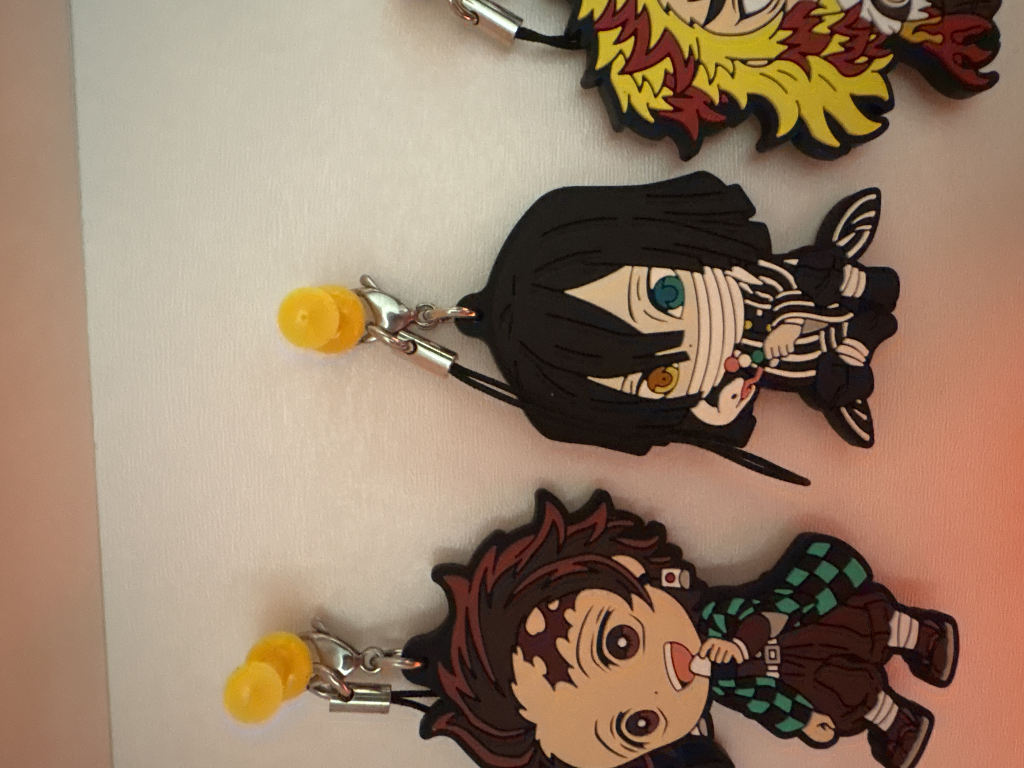 The image shows three cartoon character keychains against a white background. The keychains appear to be rubber or silicone, and each has a yellow charm and a silver-colored clasp.
From bottom to top, the characters are:
1. Tanjiro Kamado from "Demon Slayer", wearing his green and black checkered haori.
2. Obanai Iguro, also from "Demon Slayer", with bandages covering his mouth and a black and white patterned haori.
3. Kyojuro Rengoku, also from "Demon Slayer", with his distinctive flame-like hair.
There is no indication that this image was taken in Plovdiv, Bulgaria. The background is plain, and there are no identifying features related to a specific location.