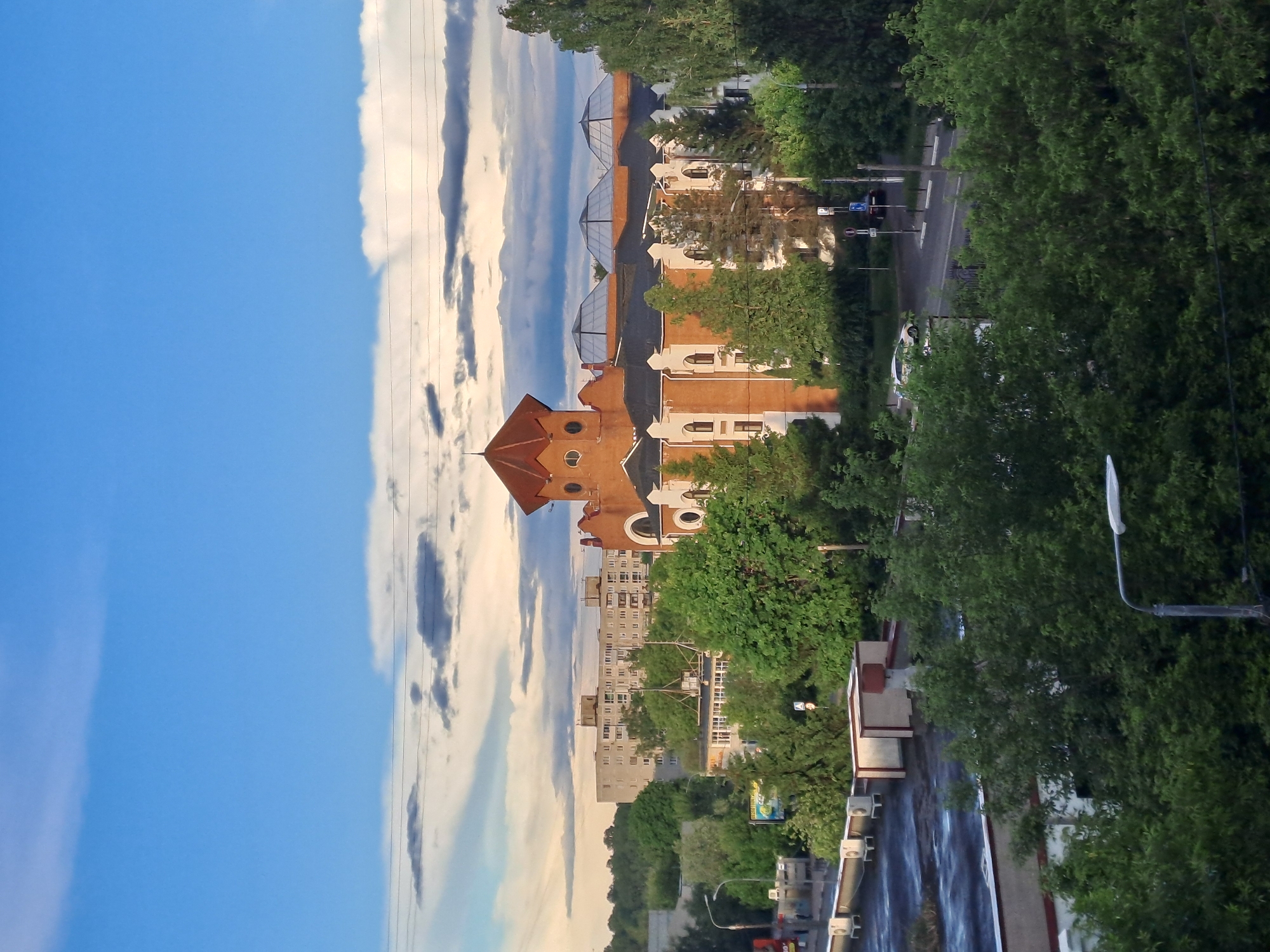 The image presents an elevated perspective of a street in Elektrostal, Russia. The scene is characterized by a mixture of architectural styles, ranging from a brick building with a prominent turret-like structure to more utilitarian apartment buildings in the background. The buildings are interspersed with mature trees, creating a green canopy that partially obscures the street below. A road runs alongside the buildings, with occasional lampposts visible. The sky is a clear blue with scattered clouds. There are vehicles on the road.