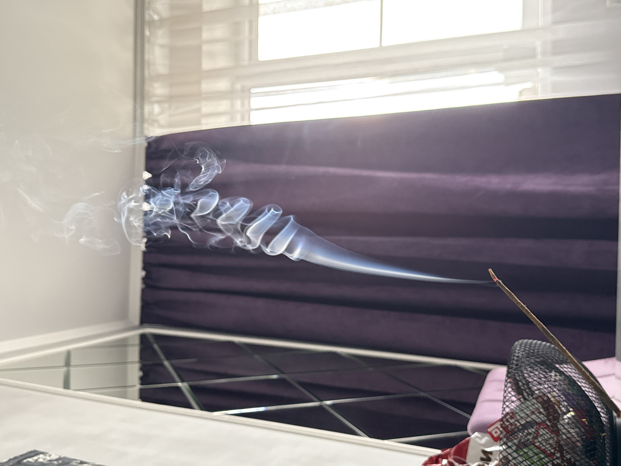 A close-up captures an incense stick burning, producing a plume of smoke that spirals and fades into the air. The stick, held in a black wire mesh container, is positioned in front of a dark purple curtain or backdrop. Light from a window with blinds illuminates the scene. The image is taken indoors, but there are no visible identifying features that would place it in a specific location.