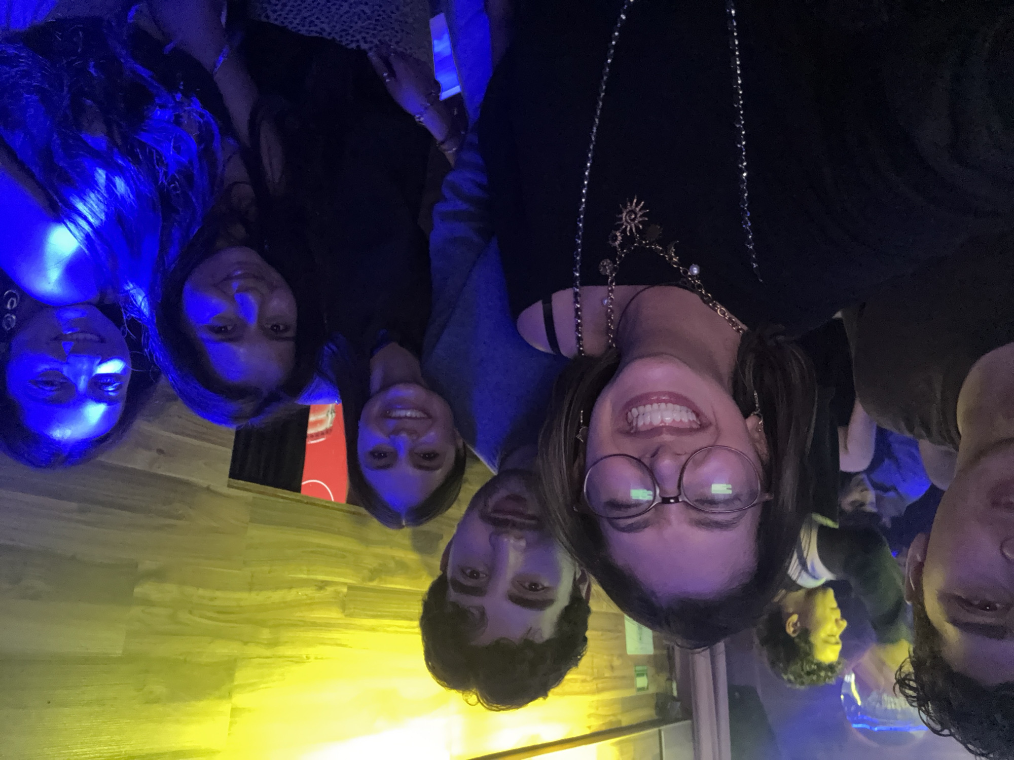A rotated selfie image shows six people under blue and yellow lighting, possibly indoors. Five are in focus and positioned closely together with one man on the right only partially in the frame. The person closest to the viewer is a smiling woman with glasses wearing a black top and jewelry. The background is not clear but seems to be a bar setting. The city of Puebla, Mexico, cannot be confirmed from the image.