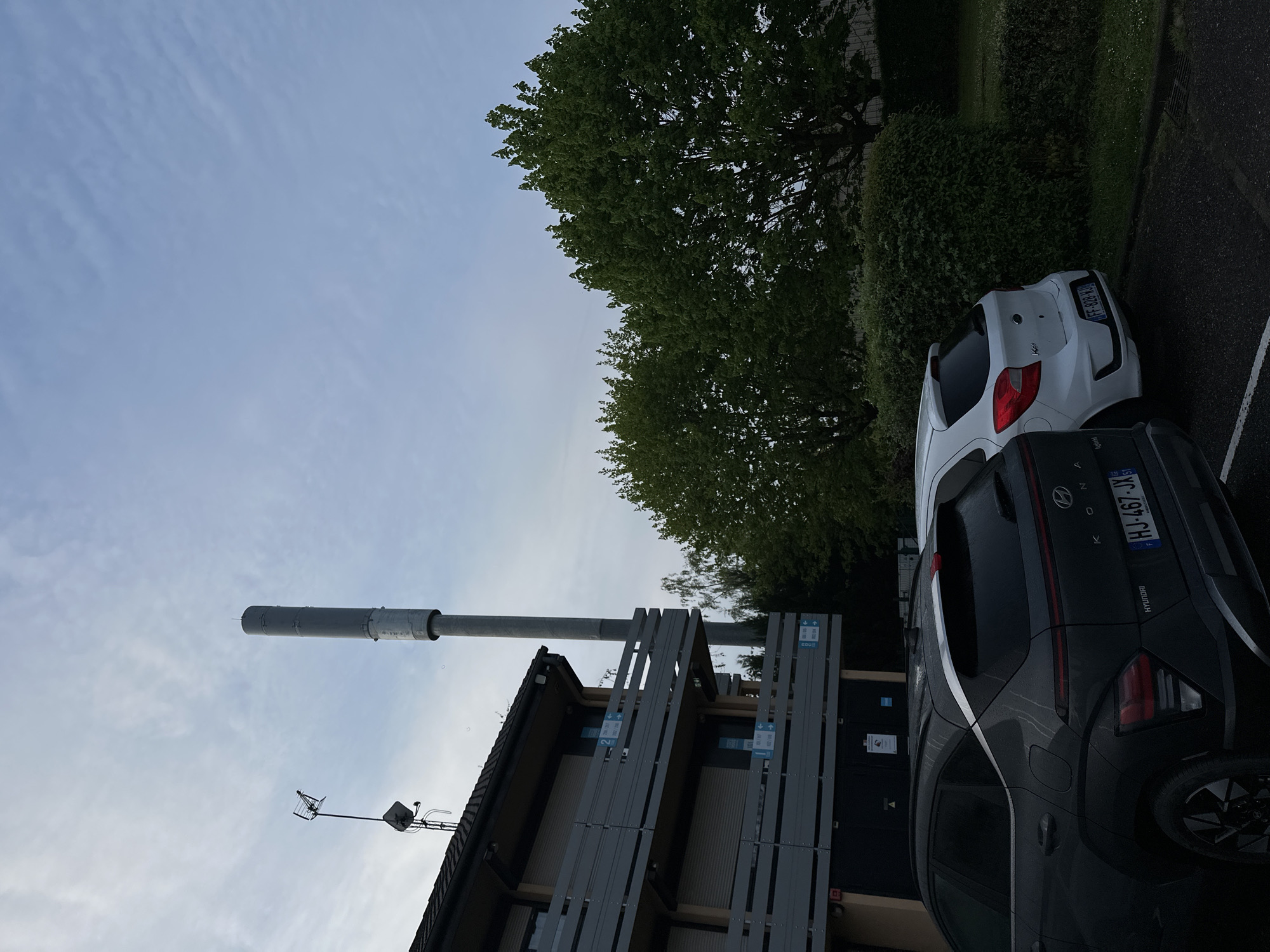 The image depicts a scene in Strasbourg, France, featuring a gray Hyundai Kona (license plate HJ-467-JX51) parked beside a white Ford Ka (license plate FF-828-NJ). Behind the cars are a tree, and a building with a tall cylindrical structure, likely a ventilation shaft or chimney, extending from the roof. A television antenna is also mounted on the roof. The building is characterized by what looks like a multi-level external access structure, possibly a staircase or walkway, with gray metal railings. The sky is overcast.