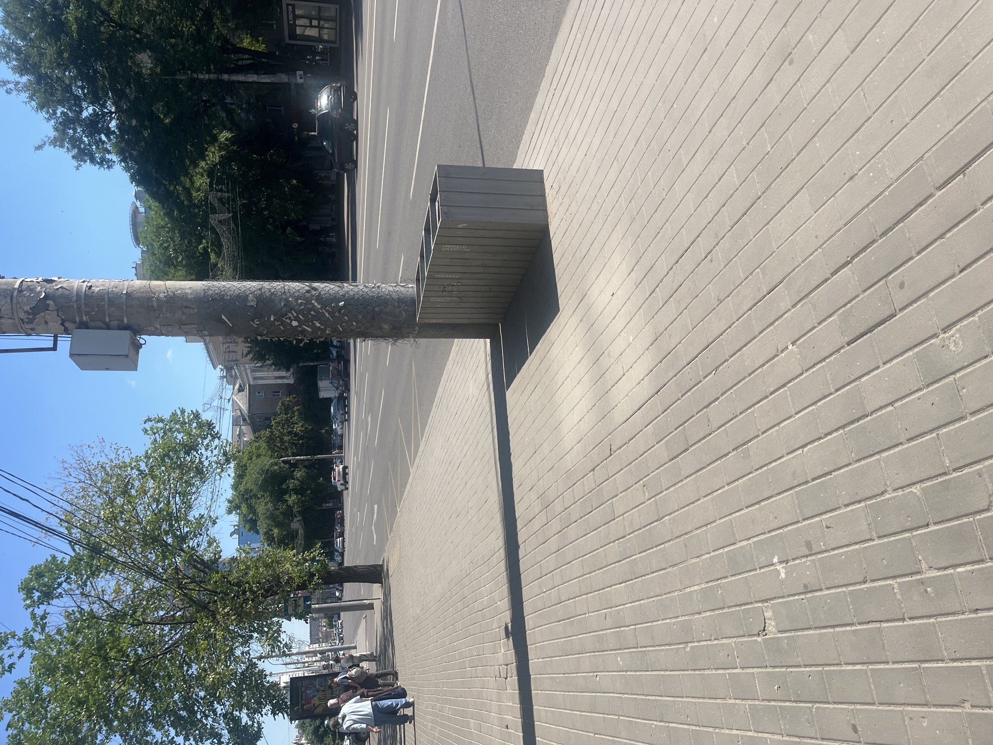 This is an outdoor, eye-level shot of a city street in Voronezh, Russia. The foreground is dominated by a paved sidewalk made of grey rectangular stones. A large, rectangular, grey planter box is positioned on the edge of the sidewalk, next to a utility pole. The utility pole is weathered and has some peeling paint, with a grey electrical box attached to it. Behind the planter box, a street with multiple lanes of asphalt is visible, with white lane markings. Several cars are driving on the street, including a dark SUV. On the far side of the street, there are buildings and trees. To the left of the planter box, on the sidewalk, a few people are standing and walking. One person in a light blue shirt and pants is walking away from the camera. Another person in a dark shirt and light pants is standing near them, facing away. A third person in a light-colored outfit is also visible. A large tree with green foliage stands on the left side of the sidewalk, casting a shadow. In the background, more trees and buildings, including a taller building with a helipad on top, can be seen under a clear blue sky.