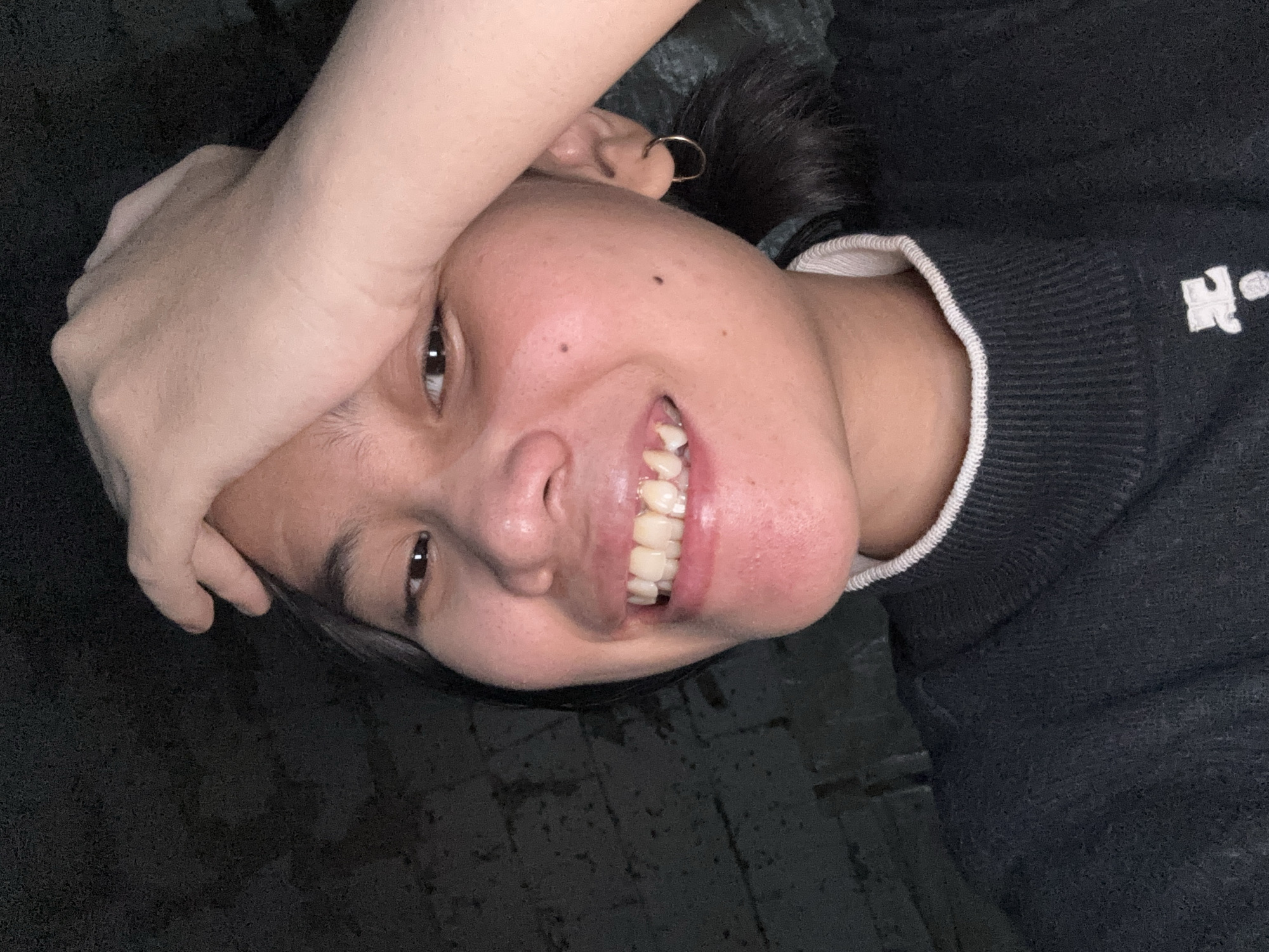 A young woman, with her hand on her forehead, smiles at the camera. She is wearing a dark sweater with a white collar. She has a gold hoop earring in her left ear. The background is dark and appears to be a brick wall.