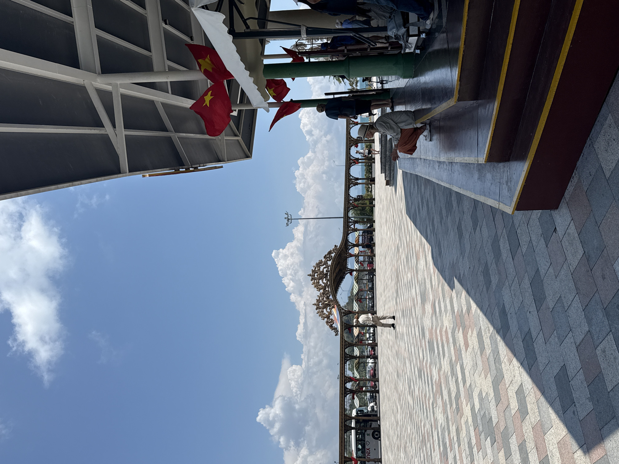 This outdoor image, taken in Nha Trang Ward, Vietnam, captures a sunny daytime scene with a distinct promenade or pier area. The perspective is slightly upward, showing a vast blue sky with scattered, fluffy white cumulus clouds.
In the immediate foreground, to the left, is a white-framed overhead canopy, likely part of a building or covered walkway. Several red flags with a yellow star, identifiable as the national flag of Vietnam, are prominently displayed, hanging from this structure. Below the canopy and to the right, a set of brown stairs with bright yellow edging leads upwards. Several people are visible under the canopy or near the stairs; one person in an orange skirt and light top is crouched near the bottom steps.
The ground is paved with a patterned mosaic of grey, light pink, and white rectangular tiles, creating a textured surface. A sharp, diagonal shadow casts across a significant portion of this tiled area on the right, indicating strong sunlight, likely from late morning or early afternoon.
Stretching across the midground is an ornate, dark-colored archway or decorative barrier, featuring intricate scrollwork. Beyond this archway, glimpses of green palm trees and distant structures can be seen, suggesting a coastal or urban setting. Several people are walking or standing along this midground area. One person, dressed in a light shirt and dark pants, is walking on the tiled path near the archway. Below the archway, several vehicles are parked; one white vehicle clearly shows the partial text "nex" or "inex" on its side.
The overall atmosphere is bright and open, indicative of a public space during good weather.
