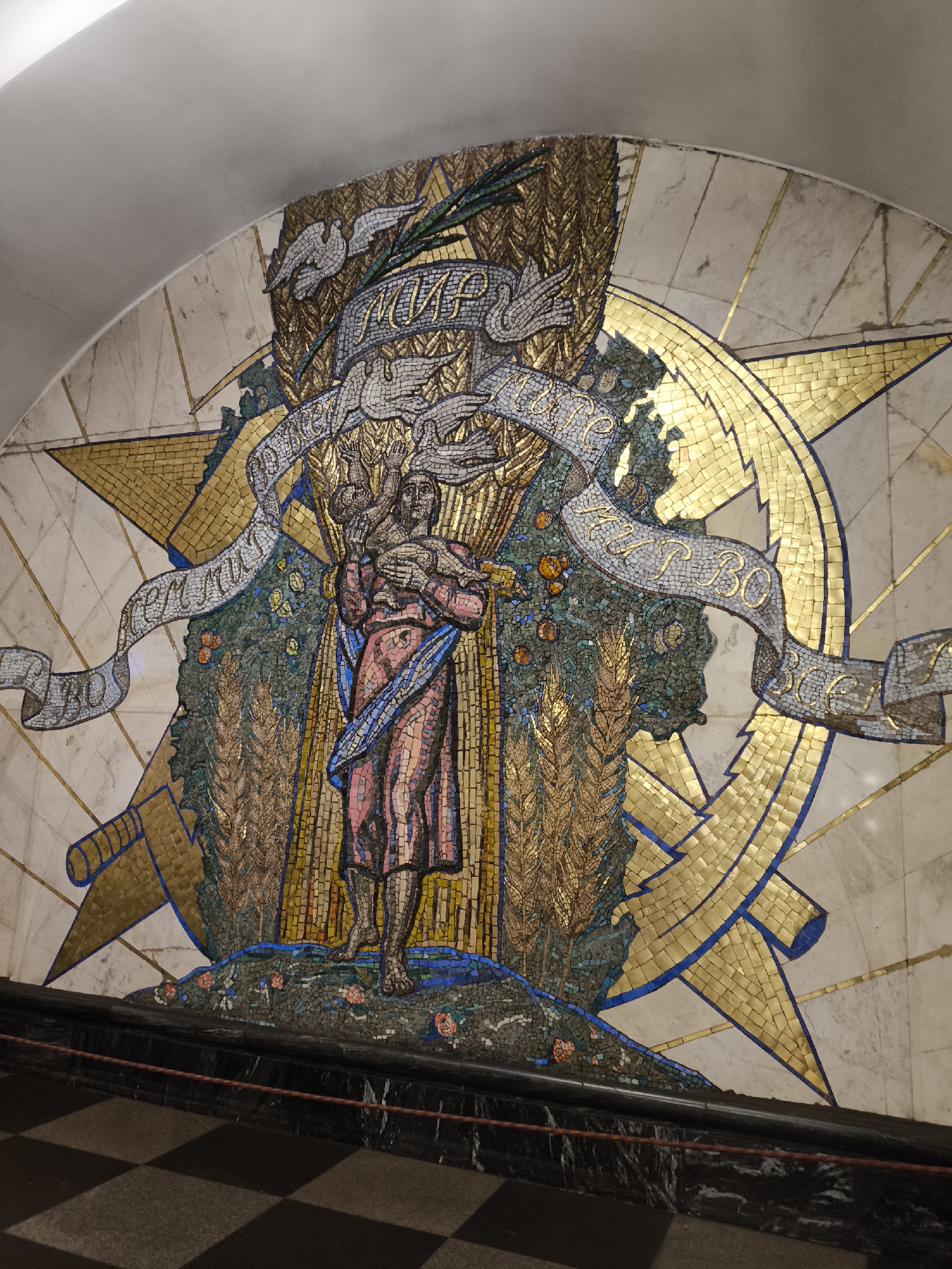 This is a mosaic artwork depicting a central female figure holding a child. She is surrounded by wheat stalks and fruit, with two doves and a laurel wreath above. A large golden star is visible behind the figure. The mosaic appears to be installed in a public space, possibly a metro station, with a curved white ceiling overhead and a checkered floor below. The inscription on the banner reads "МИР" (Peace) and "СЕМЬЯ" (Family). This mosaic is located at the Mayakovskaya metro station in Moscow, Russia.