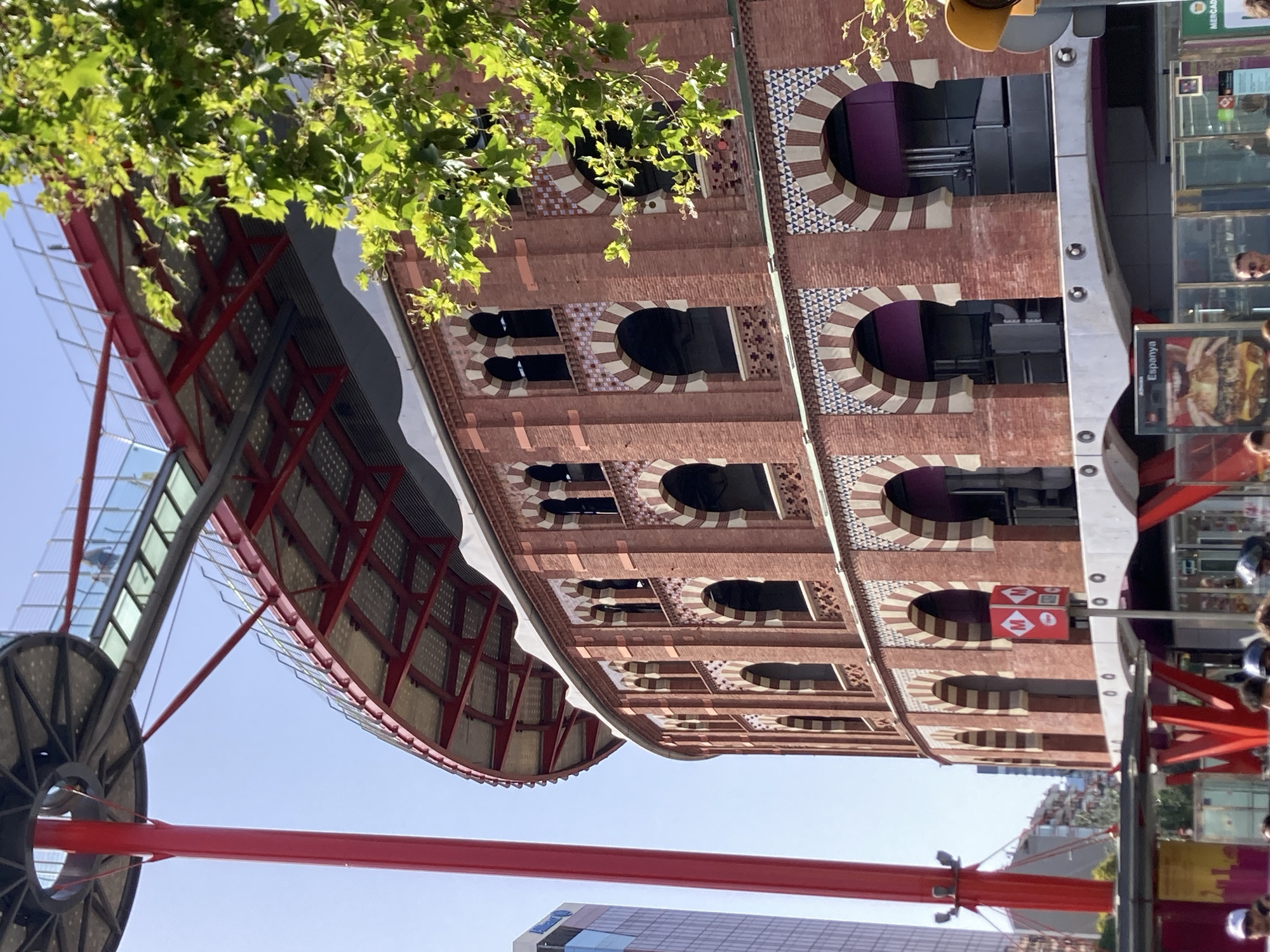 The image shows a close-up view of the exterior of the Arenas de Barcelona, a former bullring in Barcelona, Spain, that has been converted into a shopping center and leisure complex. The building is made of red brick and features a distinctive Moorish architectural style with arched windows and decorative tile work. A bright red, futuristic-looking observation deck structure extends upwards from the building, with a walkway visible above. A large green tree with vibrant leaves is in the foreground on the right, partially obscuring the building. The sky is clear and blue. In the lower portion of the image, a street-level view shows people, shop entrances, and signage, including a prominent sign for "Espanya," indicating the location is near Plaça d'Espanya.