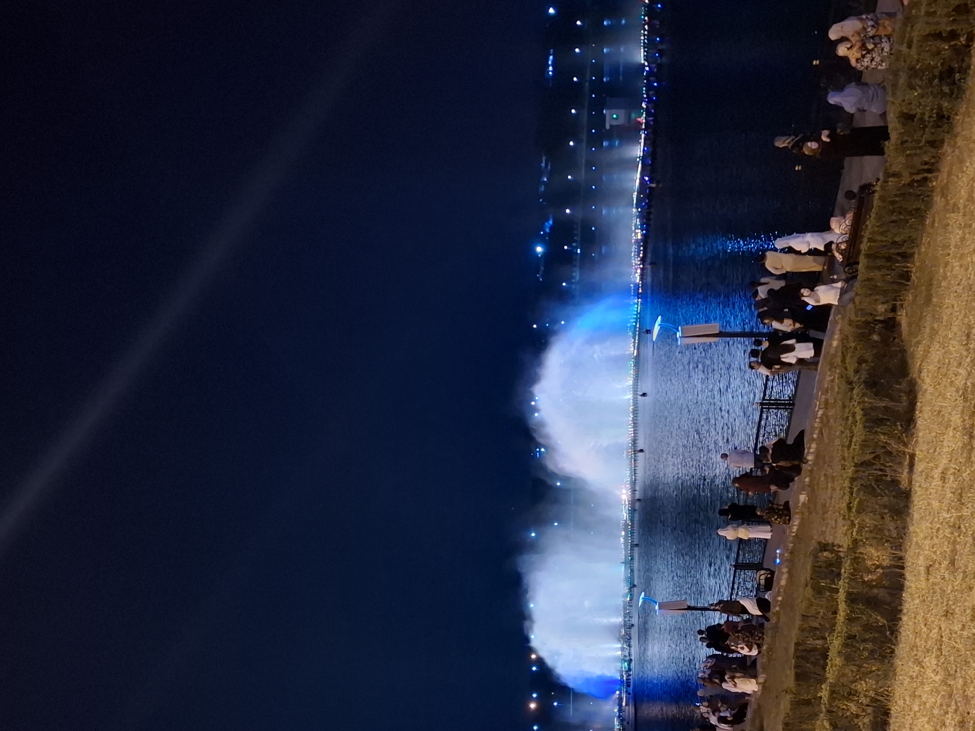 This image depicts a nighttime scene at the Grozny City Fountains in Grozny, Russia. A large, illuminated fountain display is active, projecting plumes of white and blue water into the dark sky. The fountain is situated on a body of water, with a cityscape visible in the background, dotted with numerous lights. Along the edge of the water, a crowd of people is gathered, observing the fountain show. They are standing and sitting on a walkway and a grassy embankment. Many of the individuals are wearing traditional Islamic attire, including hijabs and abayas, suggesting a diverse audience enjoying the spectacle. The lighting from the fountain and the cityscape creates a vibrant, illuminated atmosphere against the dark backdrop of the night.