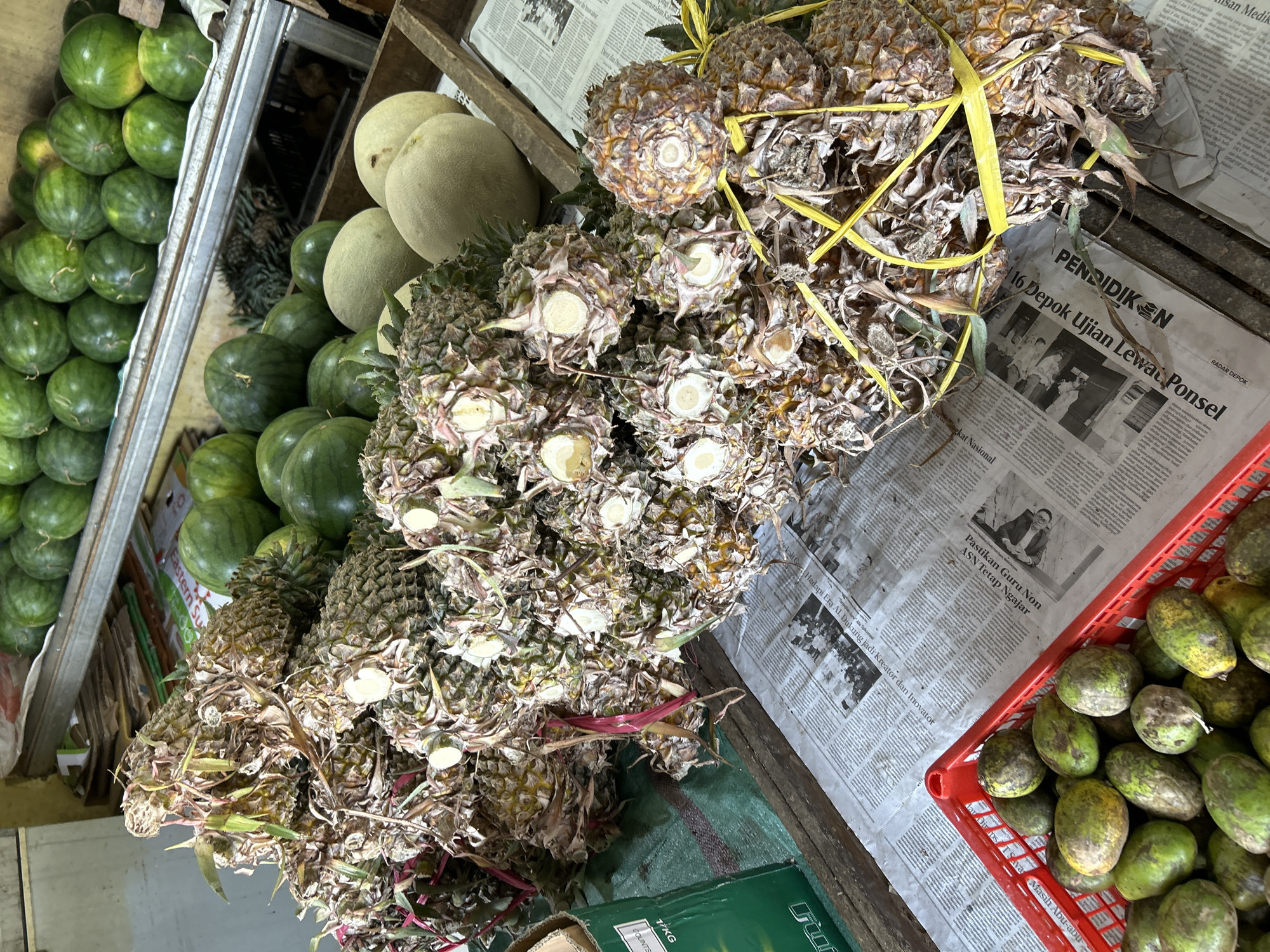 The image shows a produce stand in Depok, Indonesia, with a variety of fruits displayed. In the foreground, a red plastic basket is filled with numerous green and yellow-mottled oval fruits, likely ambarella or hog plums, resting on a layer of newspapers. Behind the basket, several piles of dried pineapples are arranged. These pineapples appear to have been harvested, with their spiky outer skins visible, and many have had their tops removed, revealing the pale, circular flesh of the core. Some of these pineapple bunches are tied together with yellow plastic strapping. In the background, more fruits are visible, including numerous small green watermelons stacked on a metal shelf. Further back, several pale, round melons, possibly honeydew or cantaloupe, are displayed in a wooden crate. The overall setting appears to be an outdoor or semi-outdoor market stall.